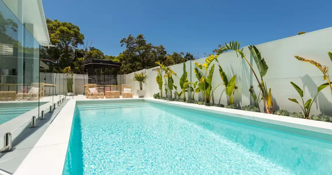 Luxury backyard swimming pool with clear blue water, surrounded by white walls and tropical plants, with lounging chairs and a glass enclosure on one side.