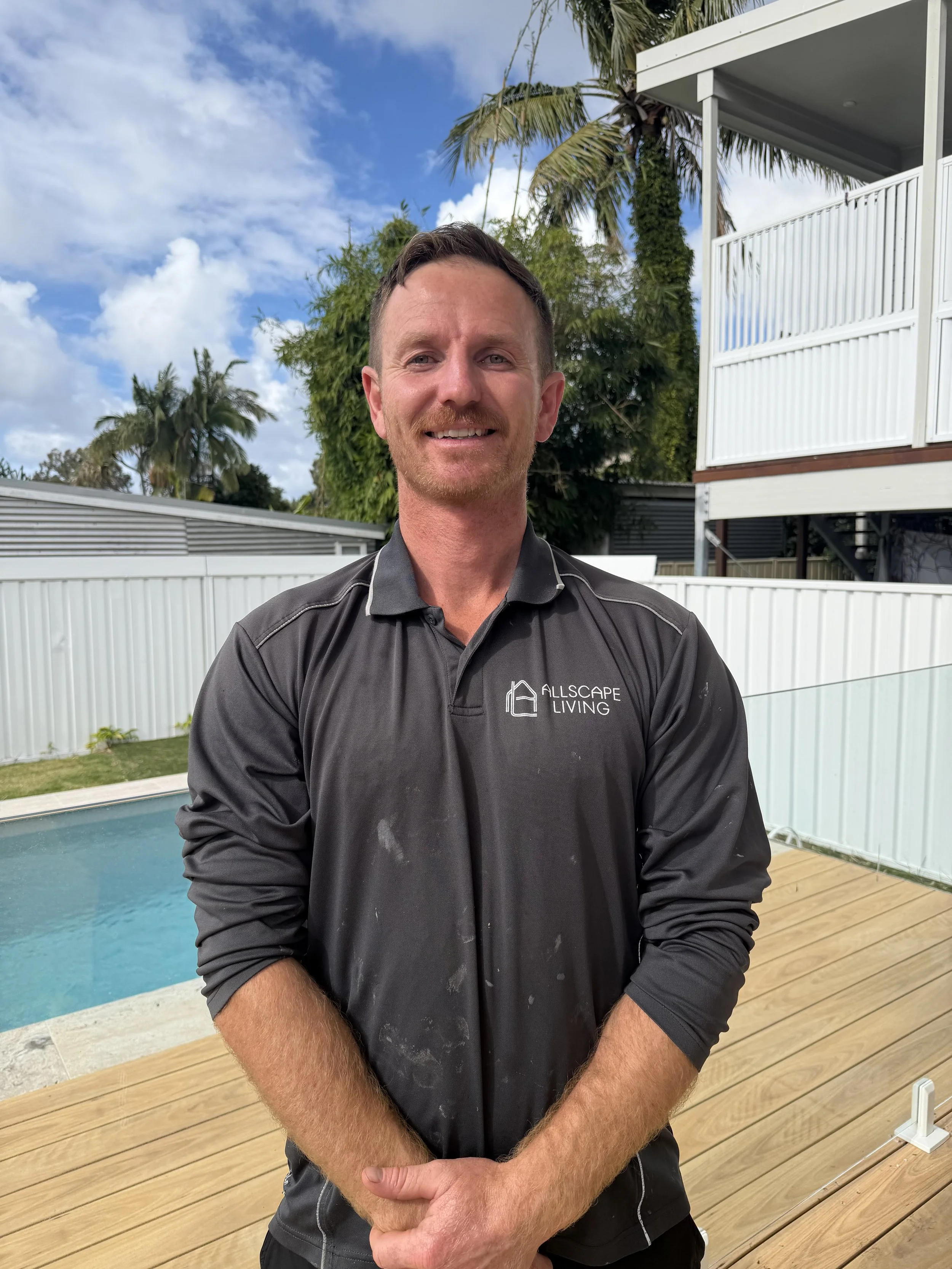 A man with light skin, short brown hair, and a reddish beard smiling in an outdoor area near a swimming pool, with a white fence, green trees, a house, and a partly cloudy sky in the background.