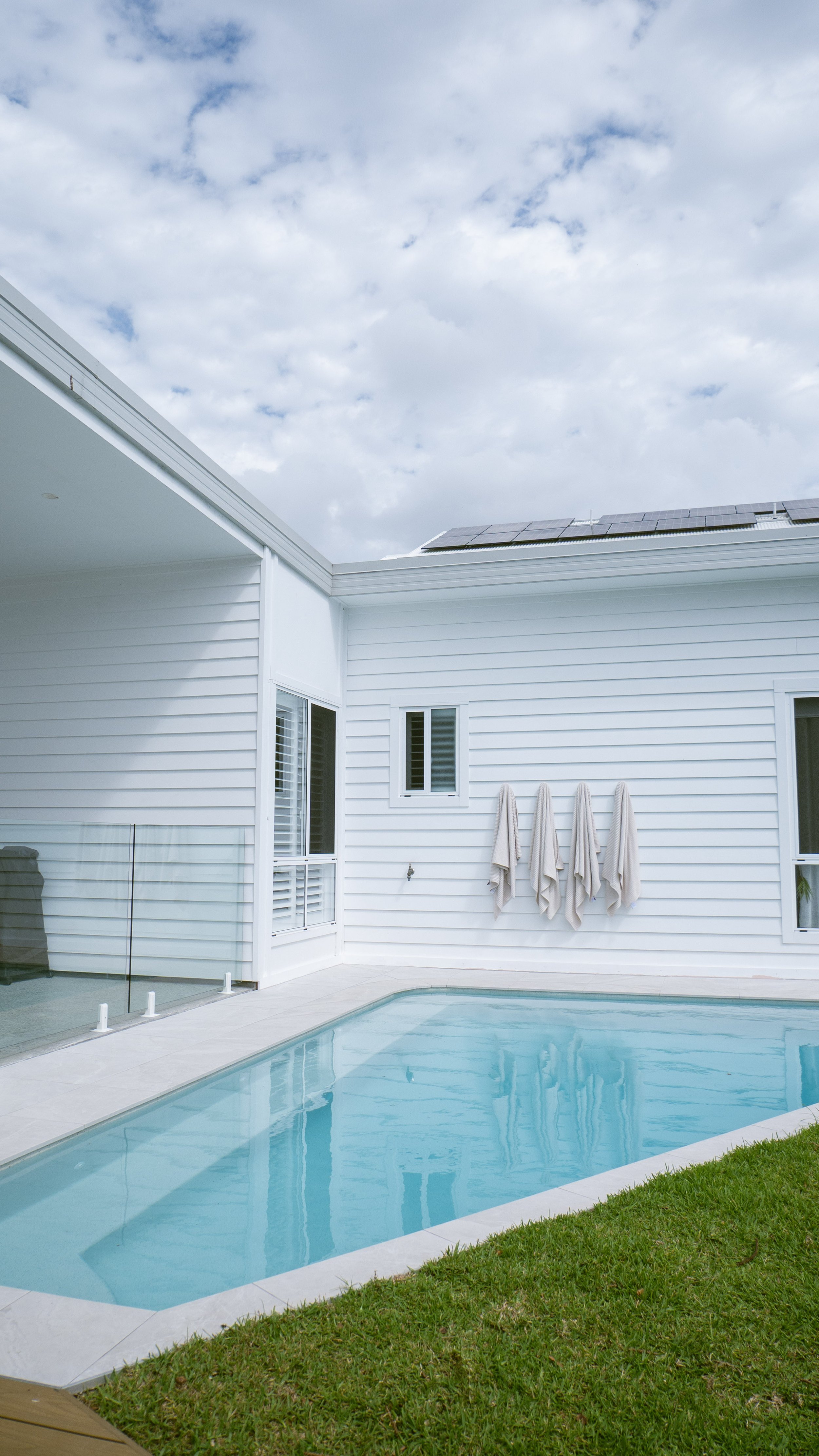 A backyard with a white house, and a swimming pool. Four towels hang on the wall, and there are solar panels on the roof, under a cloudy sky.