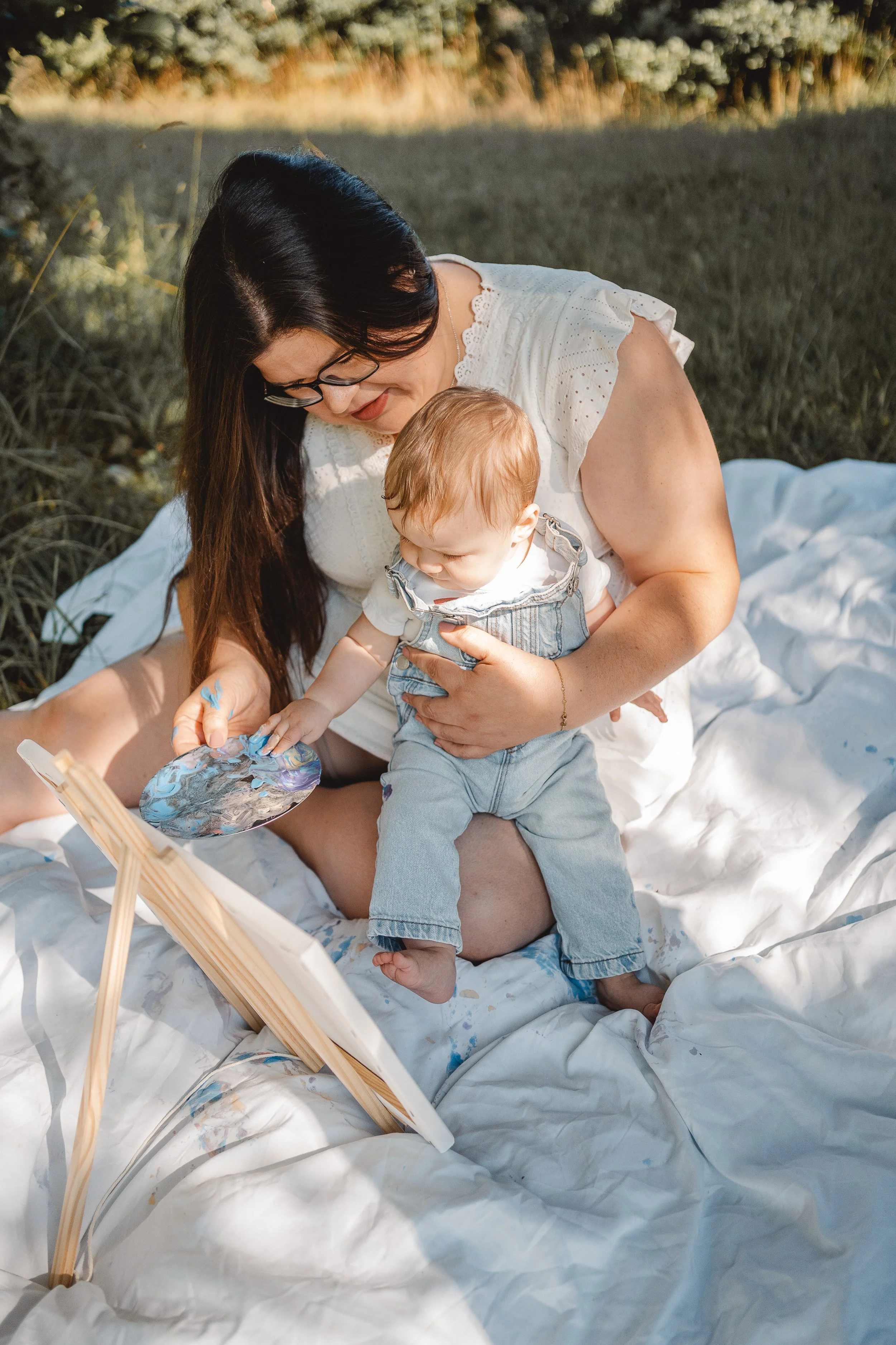 Eine Frau mit dunklen Haaren, Brille und weißem Kleid sitzt auf einer Decke im Freien und hält ein Baby im Arm. Sie spielt mit einem Maler-Set, während das Baby auf ihrem Schoß sitzt und nach den Farben greift.