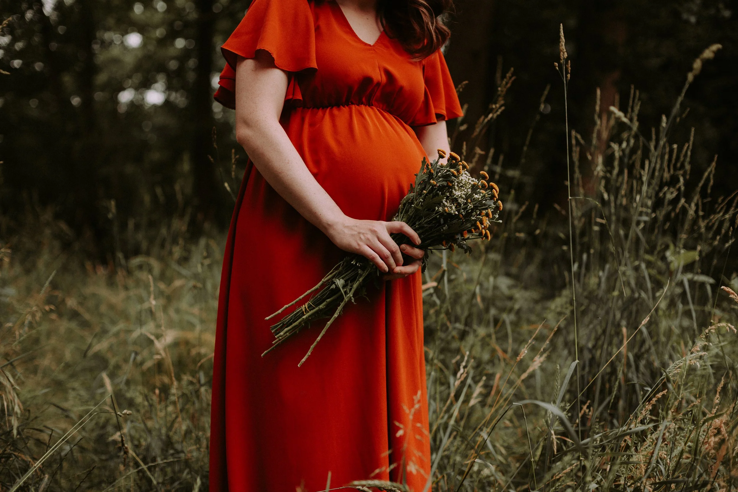 Eine schwangere Frau in einem roten Kleid hält einen Blumenstrauß in einer abgelenkten, natürlichen Umgebung.