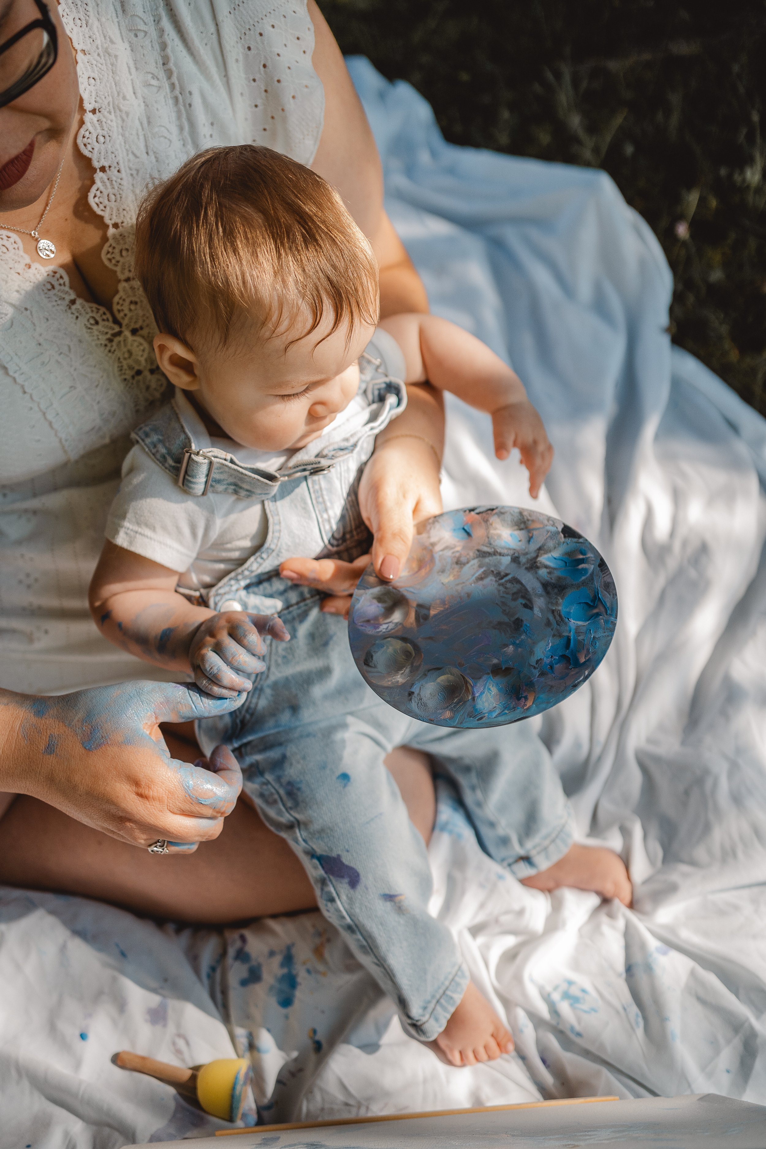 Eine Frau hält ein Baby, das mit blauer Farbe bemalt ist, auf ihrem Schoß. Das Baby trägt eine Collegejacke und sitzt auf weißem Stoff, der mit blauer Farbe bemalt ist.
