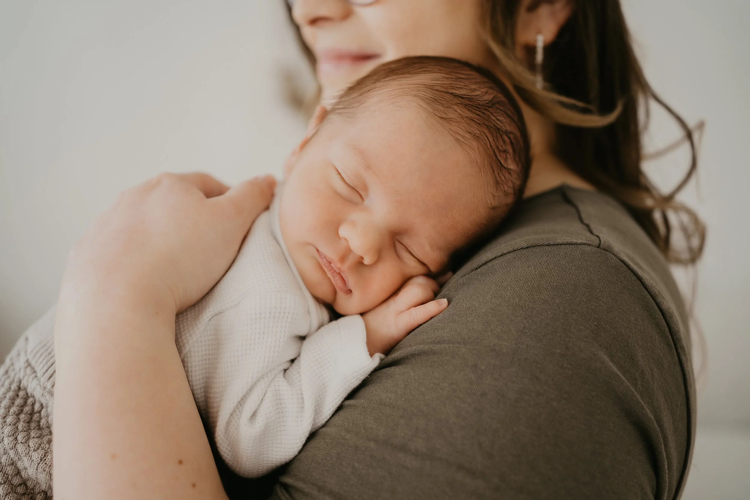 Eine Frau hält ein schlafendes Baby an ihrer Schulter. Das Baby lächelt friedlich und hat dunkles, dünnes Haar. Newbornshooting, Neugeborenenfotograf, Fotostudio Pressath