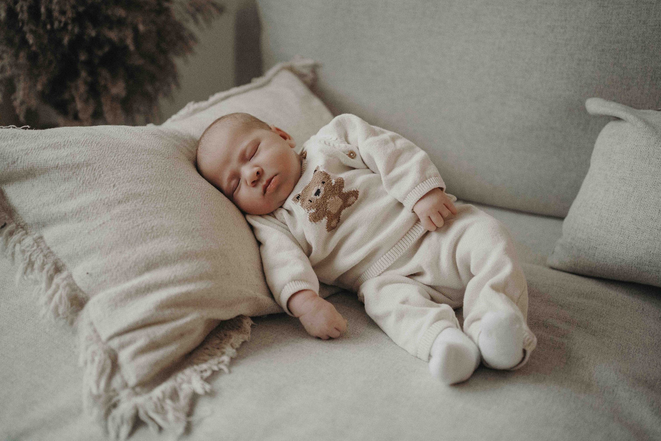 Ein schlafender Säugling liegt auf einem weichen Kissen auf einem Sofa, eingekleidet in einen beige-weißen Einteiler mit einem Bärenmuster.