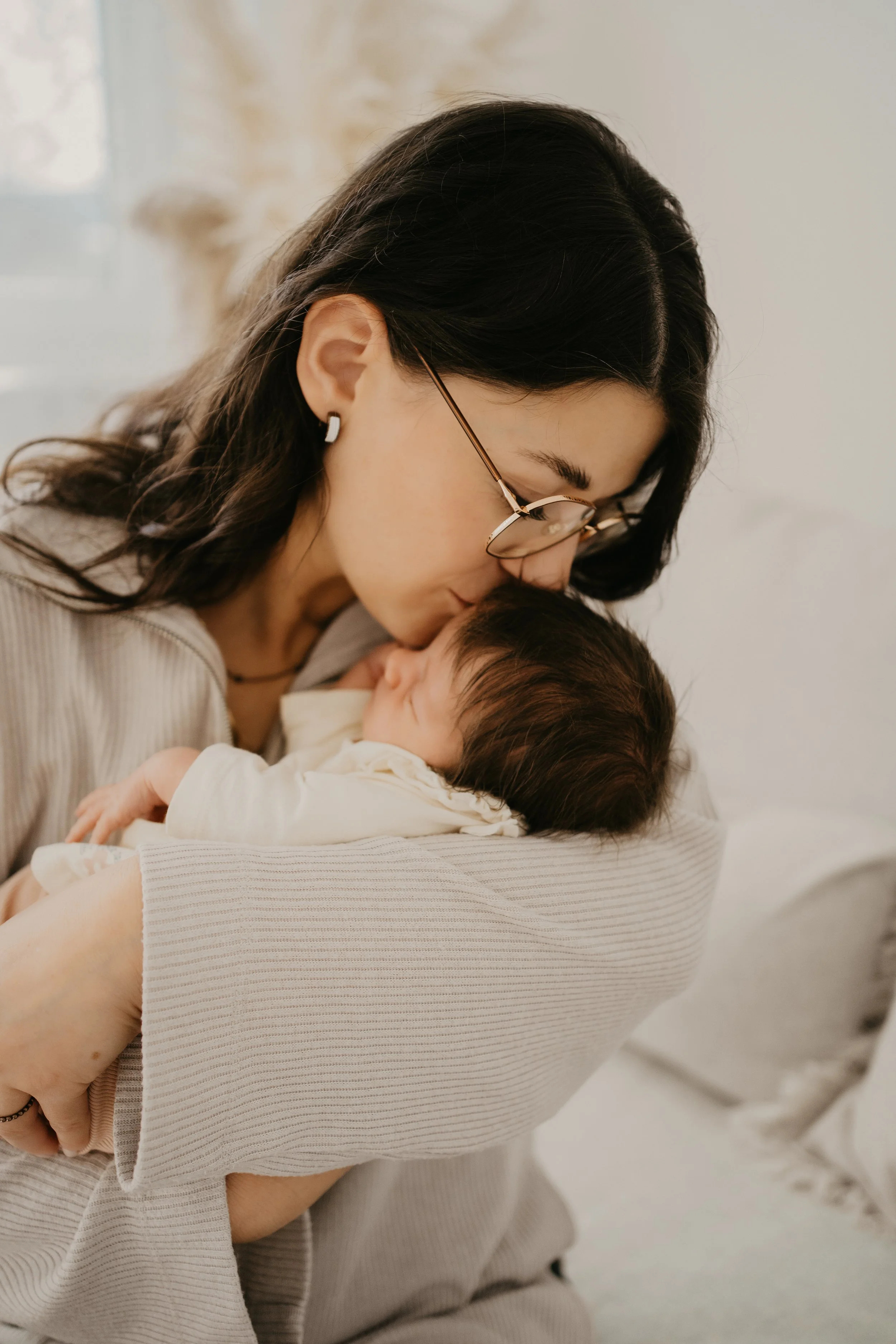 Eine Frau hält ein kleines Baby im Arm und küsst es auf die Stirn in einem hellen Raum.