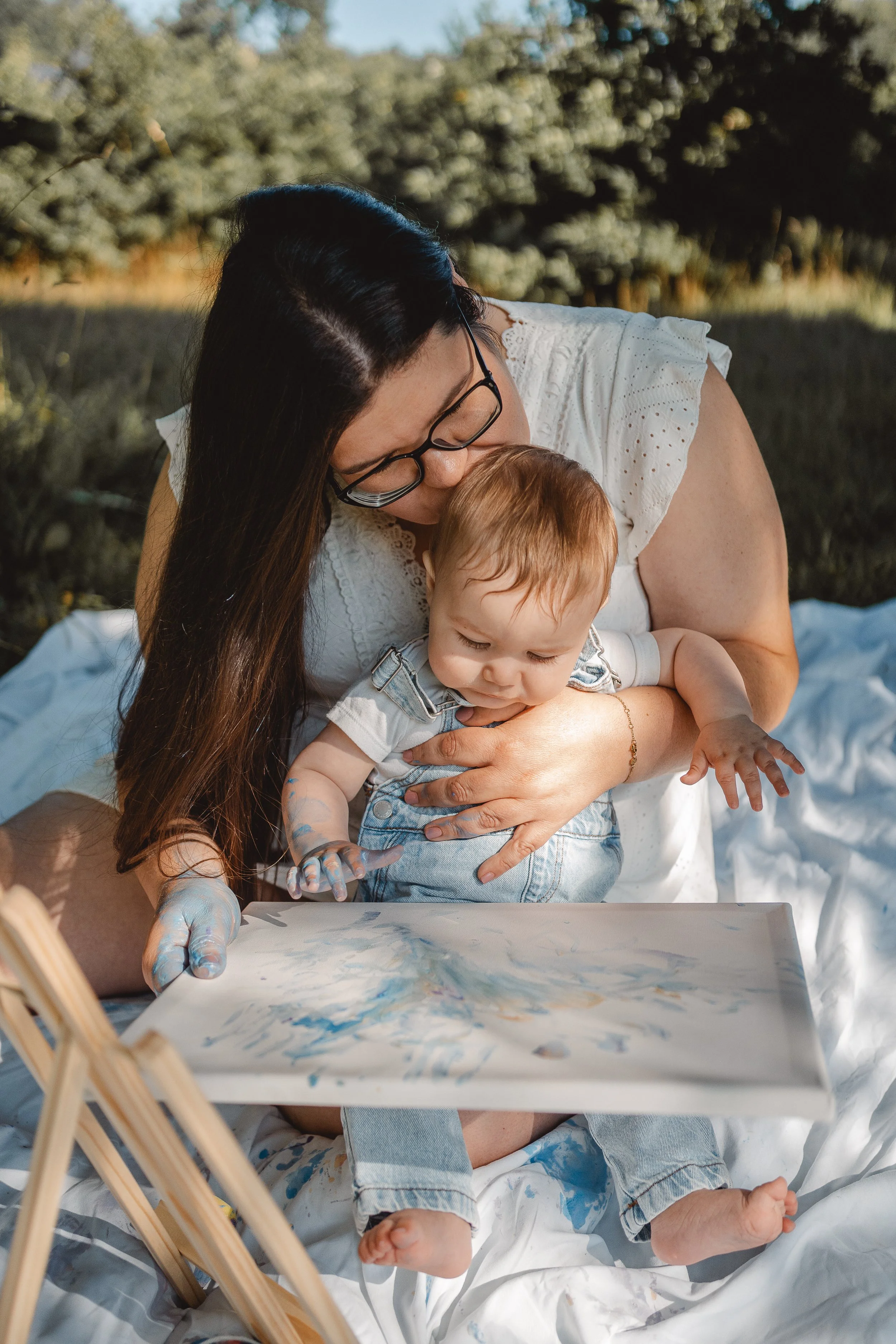 Eine Frau mit schwarzen Haaren und Brille umarmt und küsst ein Baby mit roten Haaren, das auf einem großen weißen Papier malt, im Freien bei Tageslicht.