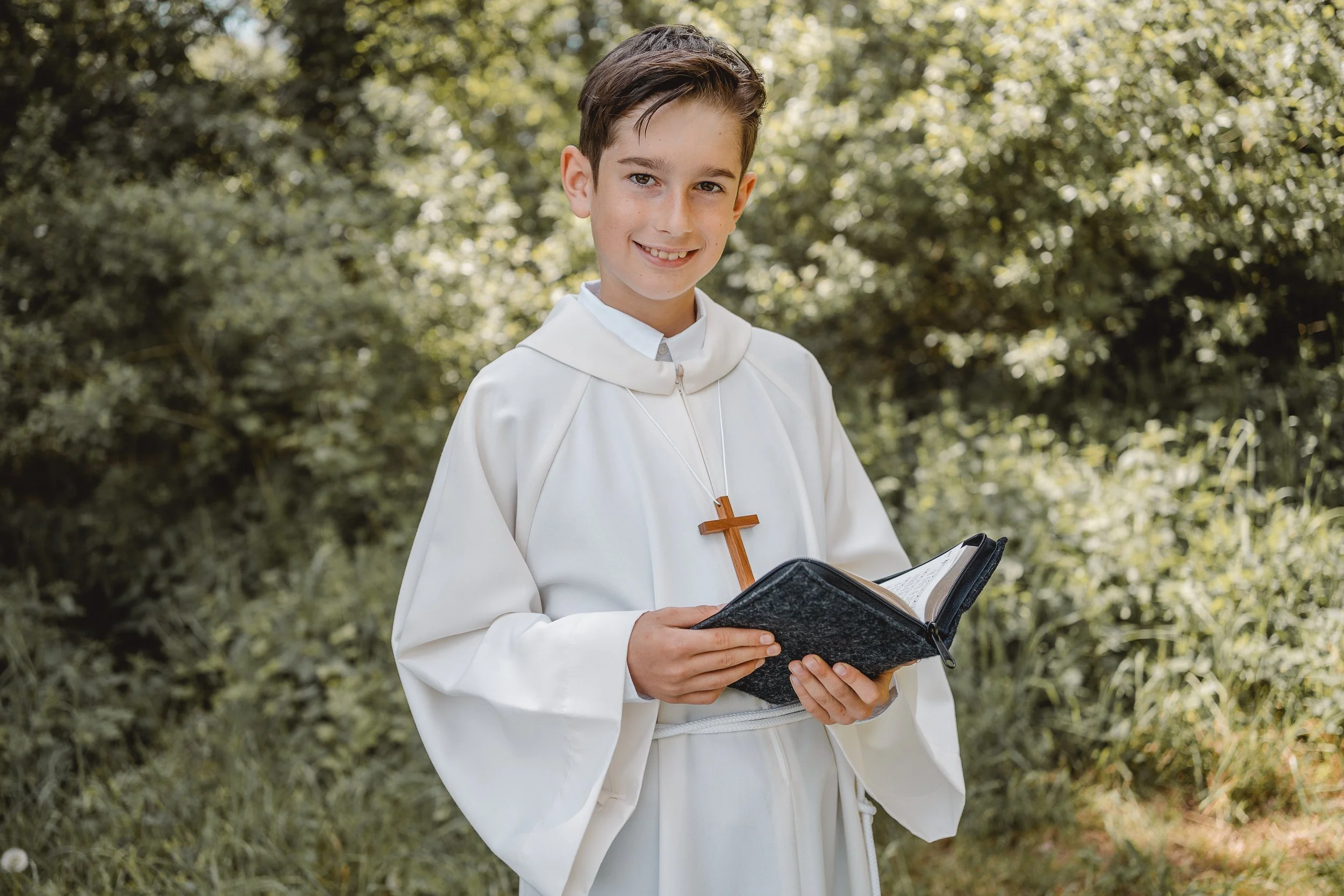 Ein junger Junge in einer weißen Kirche-Kleidung hält eine offene Bibel und lächelt. Er trägt ein Holzkreuz um den Hals. Der Hintergrund ist ein bewaldeter Bereich mit Sonnenlicht, der eine ruhige und friedliche Atmosphäre schafft.