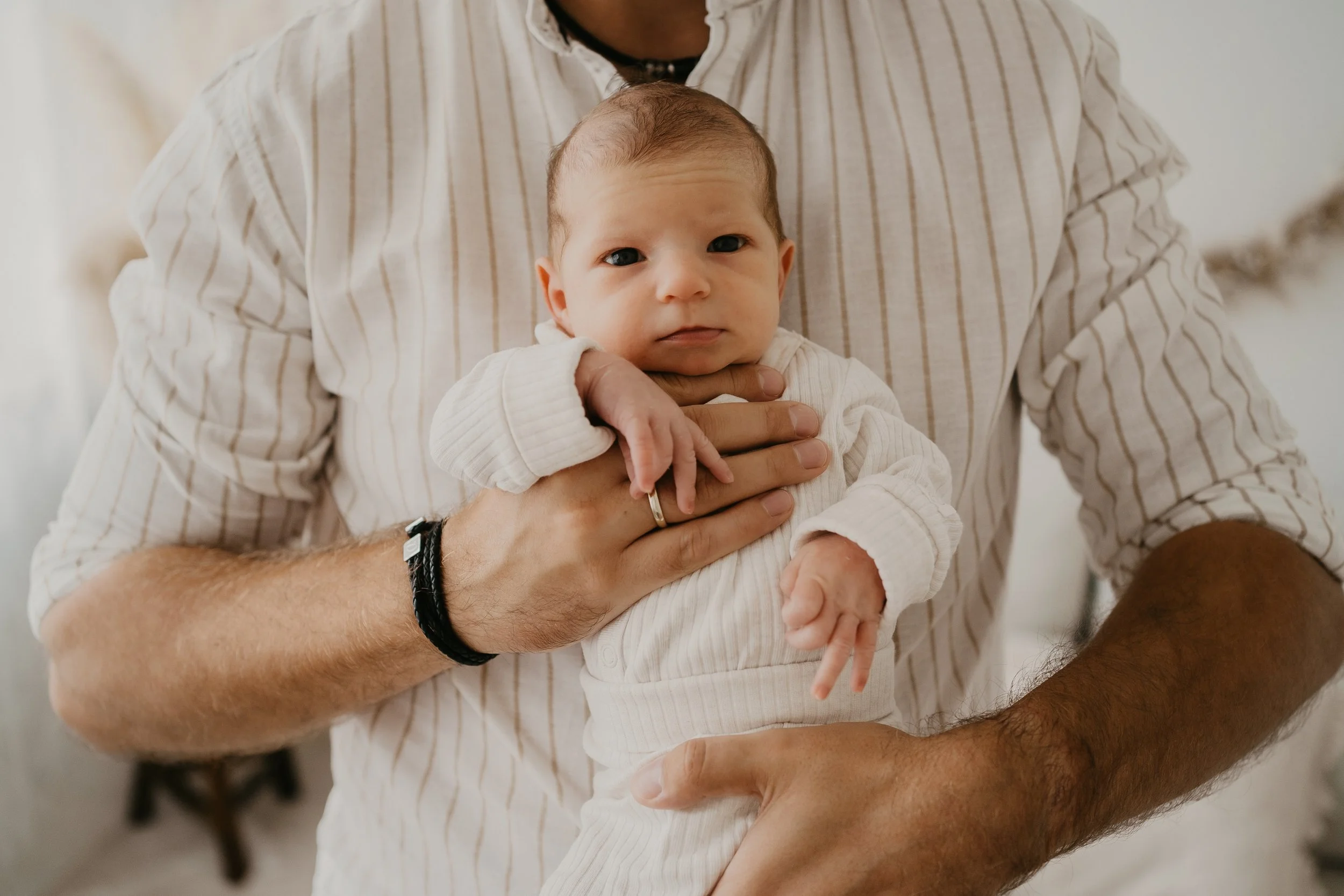 Ein Baby wird von einer Person gehalten, die einen cremefarbenen, gestreiften Hemd trägt. Das Baby trägt einen weißen Strickpullover und schaut direkt in die Kamera.