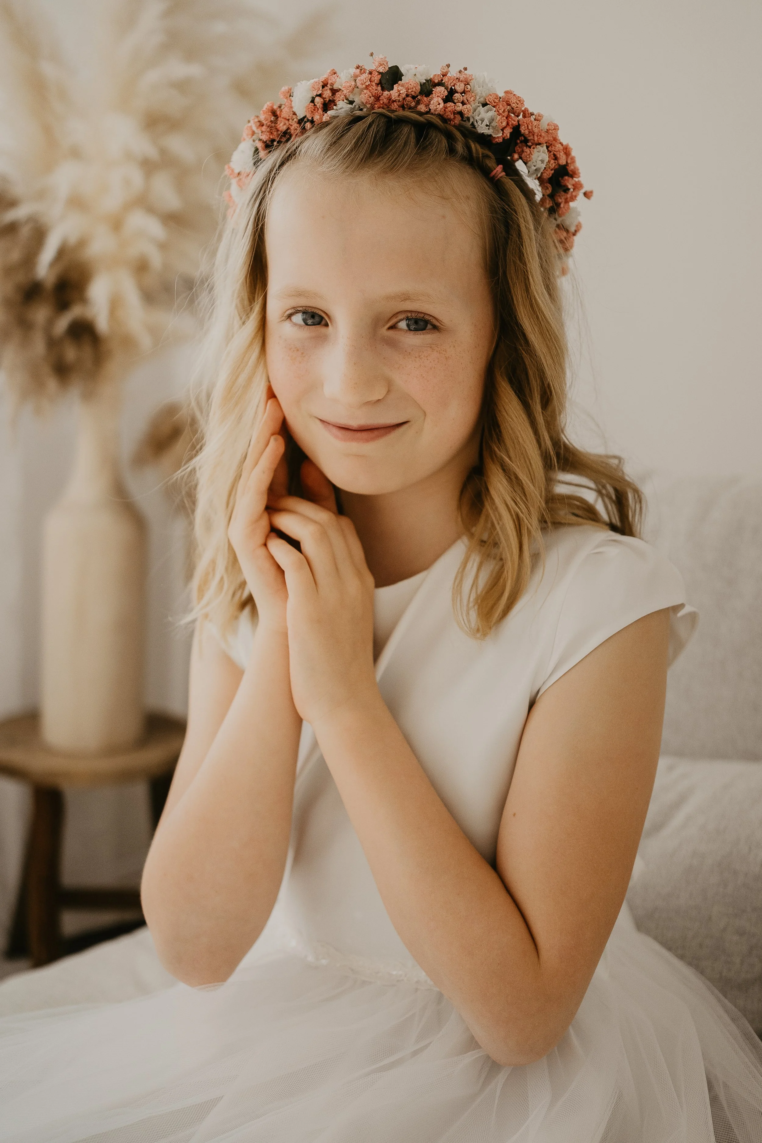 Ein junges Mädchen mit blonden, lockigen Haaren, trägt ein weißes Kleid und einen Blumenkranz auf dem Kopf, sitzt auf einem Sofa und lächelt in die Kamera.