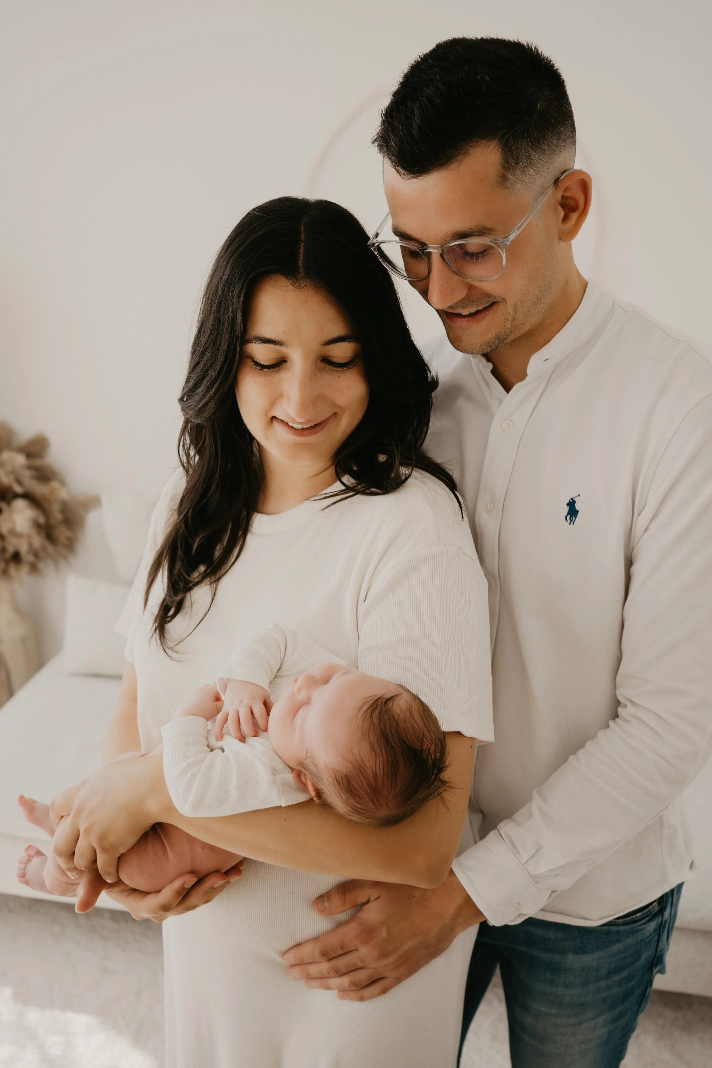Eine junge Frau hält ein Baby im Arm, während ein Mann neben ihr steht und lächelt. Die Szene ist in einem hellen Raum mit neutraler Wand im Hintergrund.