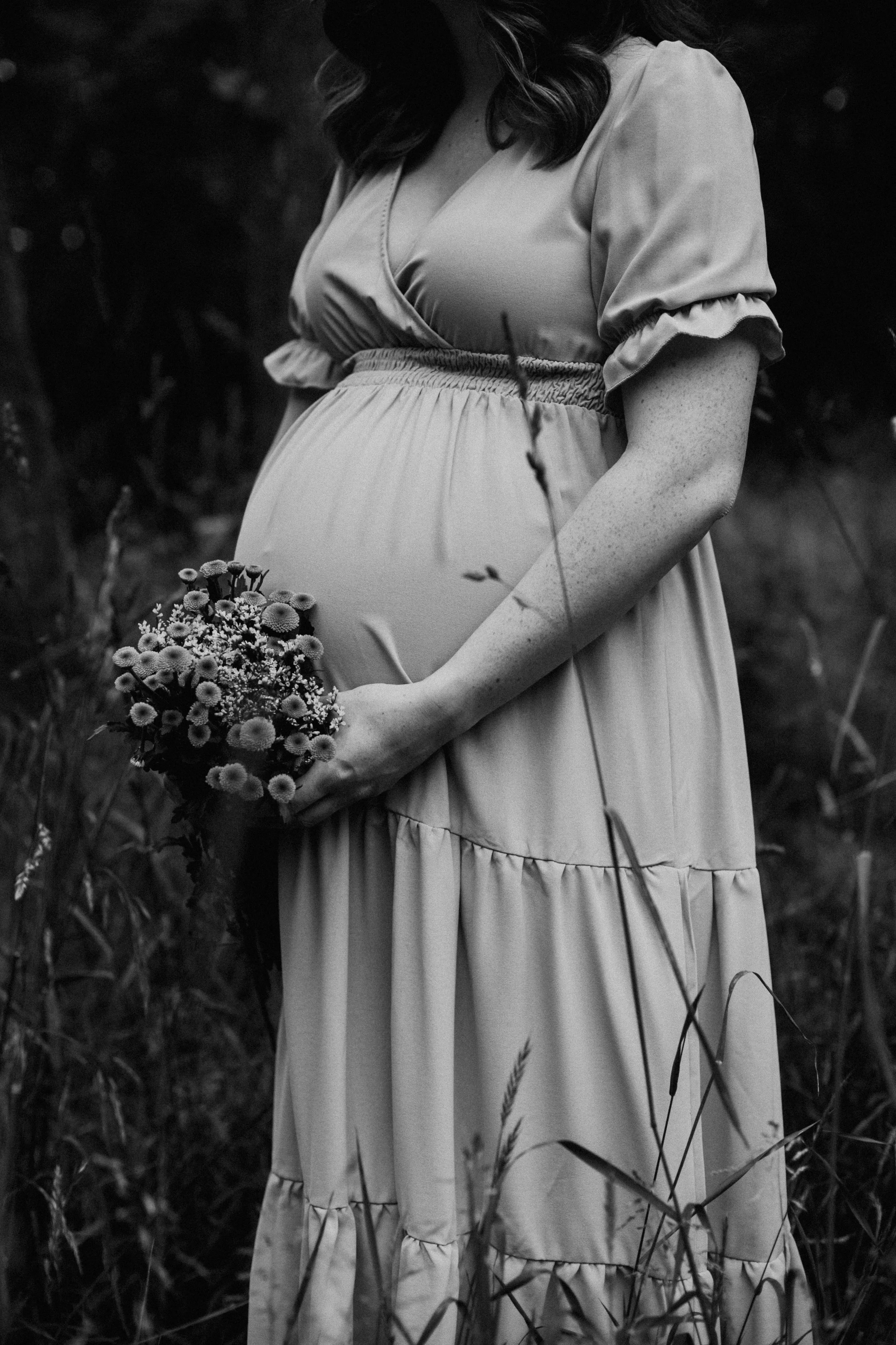 Schwarz-weiß Foto einer schwangeren Frau, die ein Kleid trägt und einen Blumenstrauß hält, im Freien in einem natürlichen Umfeld.