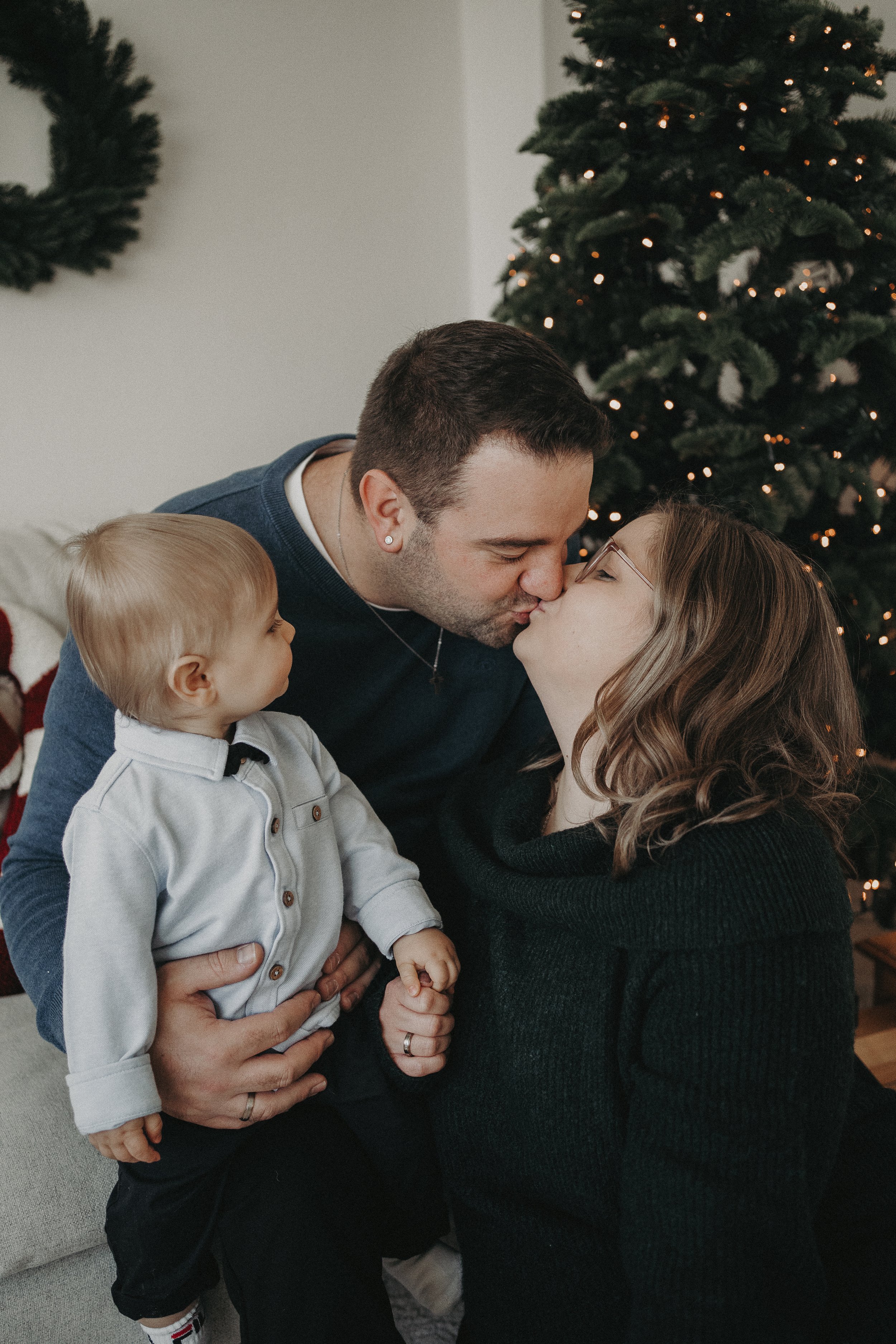 Familie bei einer Weihnachtsfeier, sie küssen sich vor einem geschmückten Weihnachtsbaum.