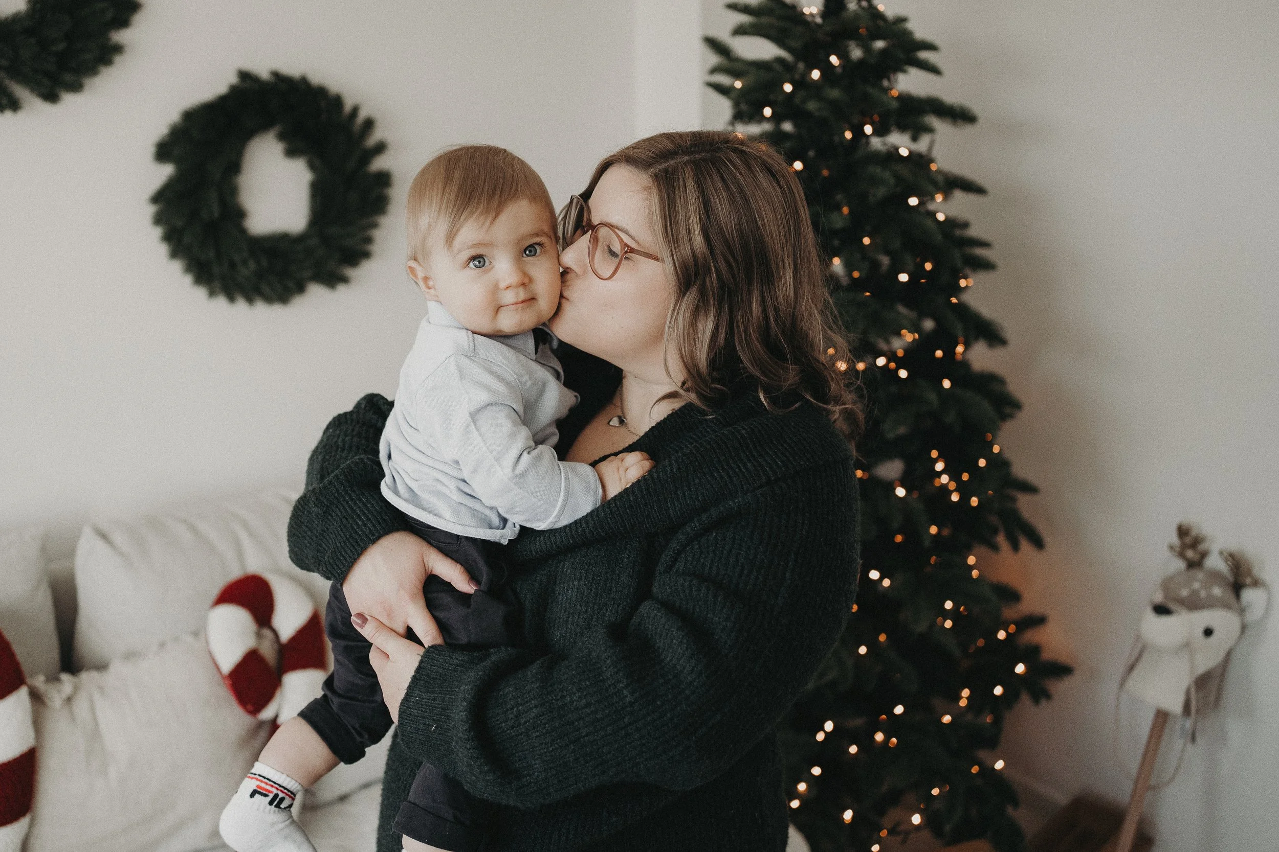 Eine Frau umarmt und küsst einen kleinen Jungen vor einem Weihnachtsbaum mit Lichtern, im Hintergrund eine Wandleiste und Dekorationen.