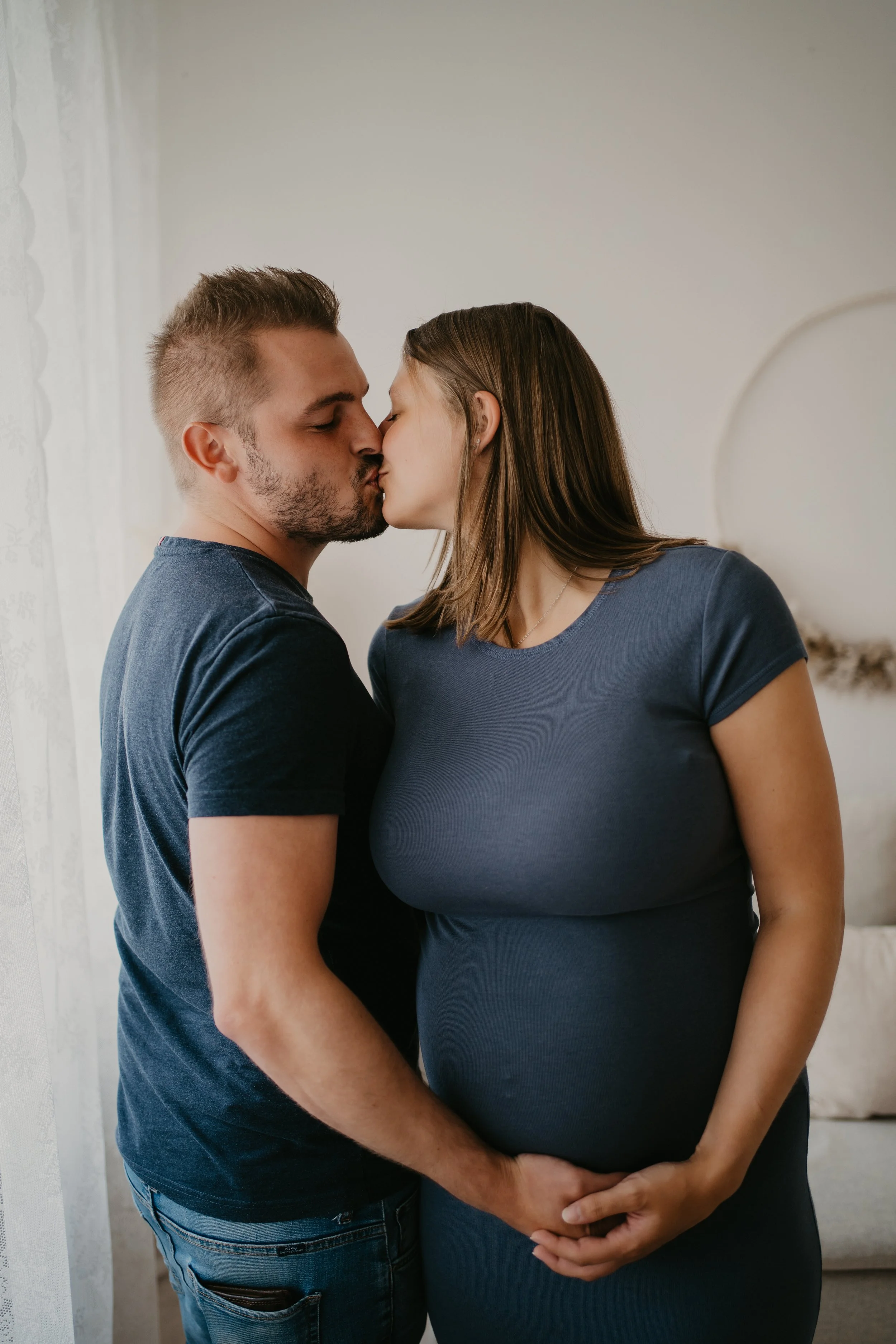 Ein Paar küsst sich liebevoll, die schwangere Frau hält ihre Babybauch mit beiden Händen, der Mann umarmt sie.