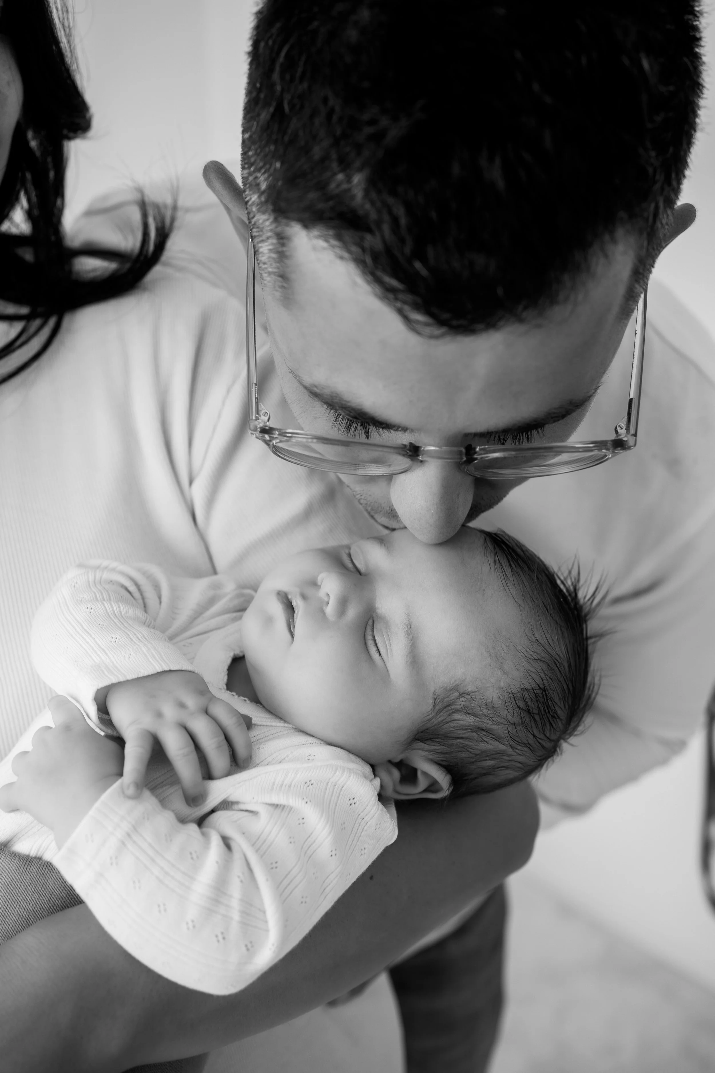 Ein Mann mit kurzen dunklen Haaren und Brille hält ein schlafendes Baby im Arm, beide in Schwarz-Weiß-Fotografie festgehalten.