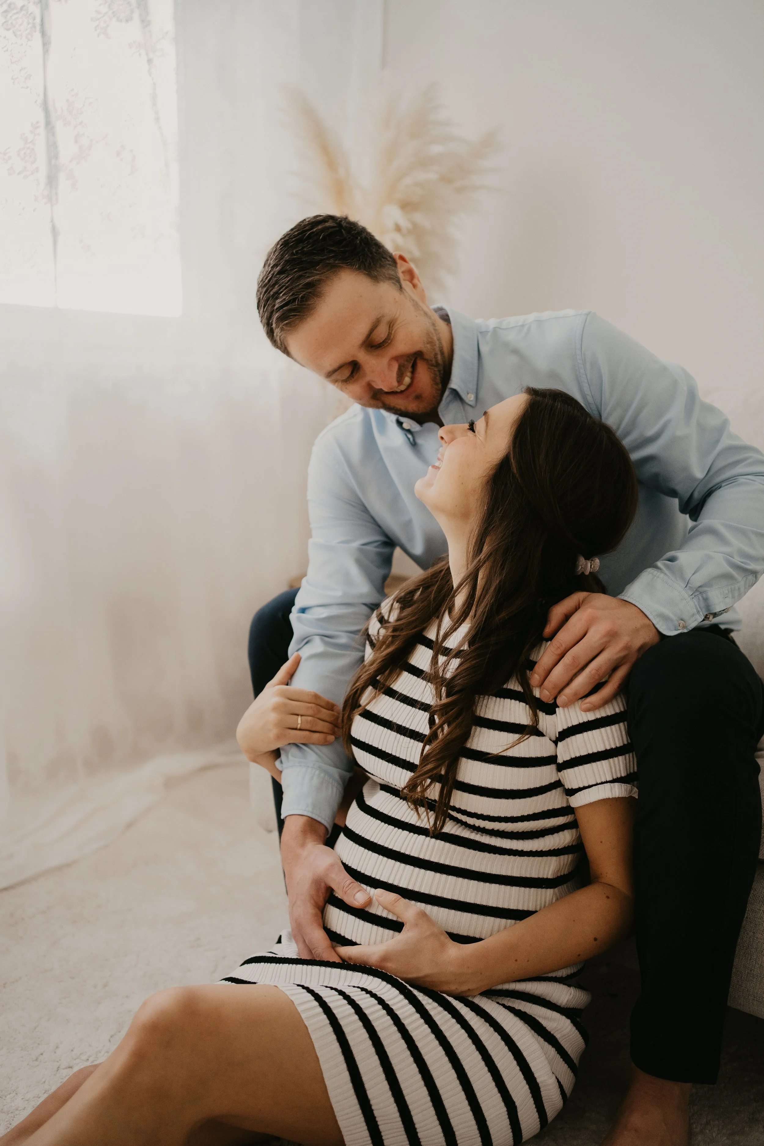 Ein glückliches Paar beim Babybauch-Posieren in einem hellen Raum, das Paar zeigt Liebe und Verbundenheit.
