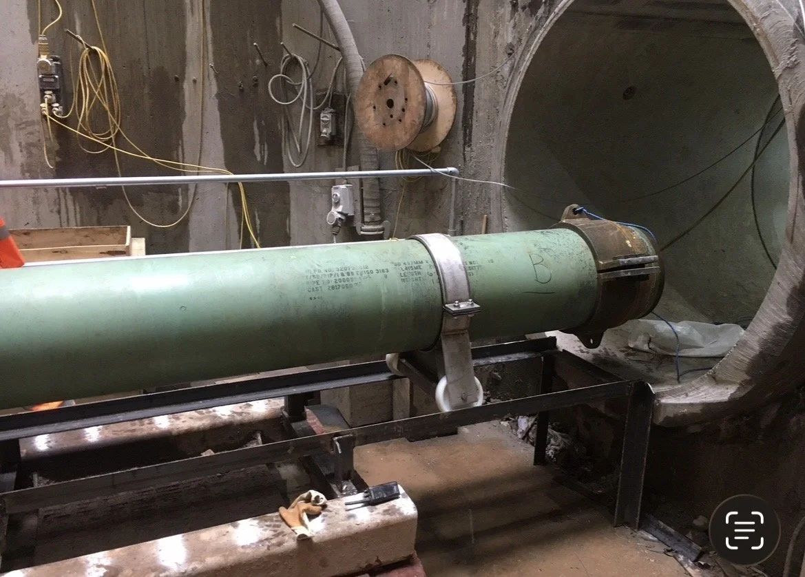Construction site showing a large green pipe mounted on a metal frame, with a tunnel opening behind it. Electrical wires and construction tools are visible around the area.
