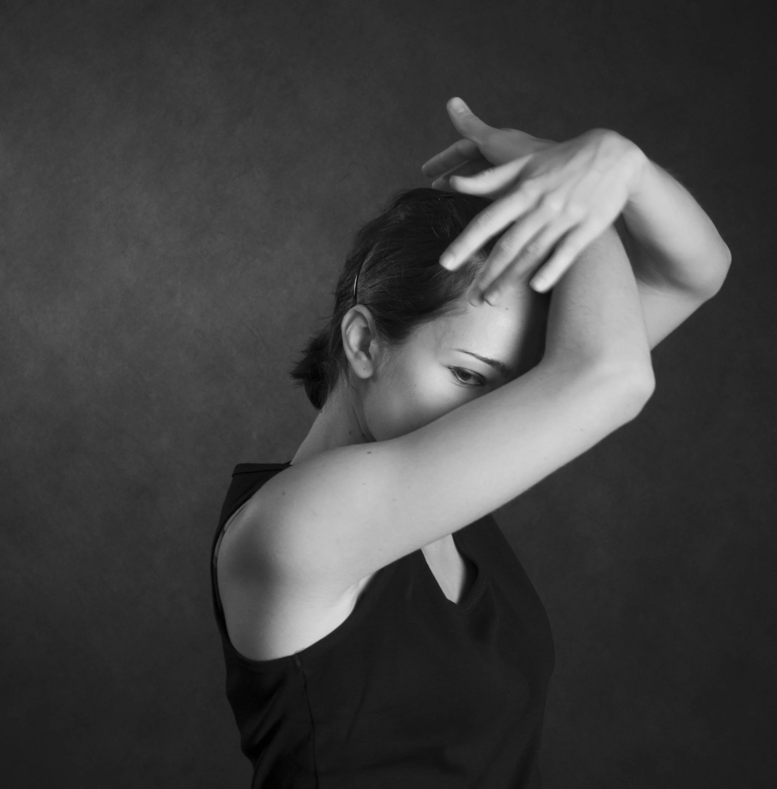 Black and white photo of a person with short dark hair, accessorized with glasses, raising their arms and touching their forehead in a pose against a dark background.