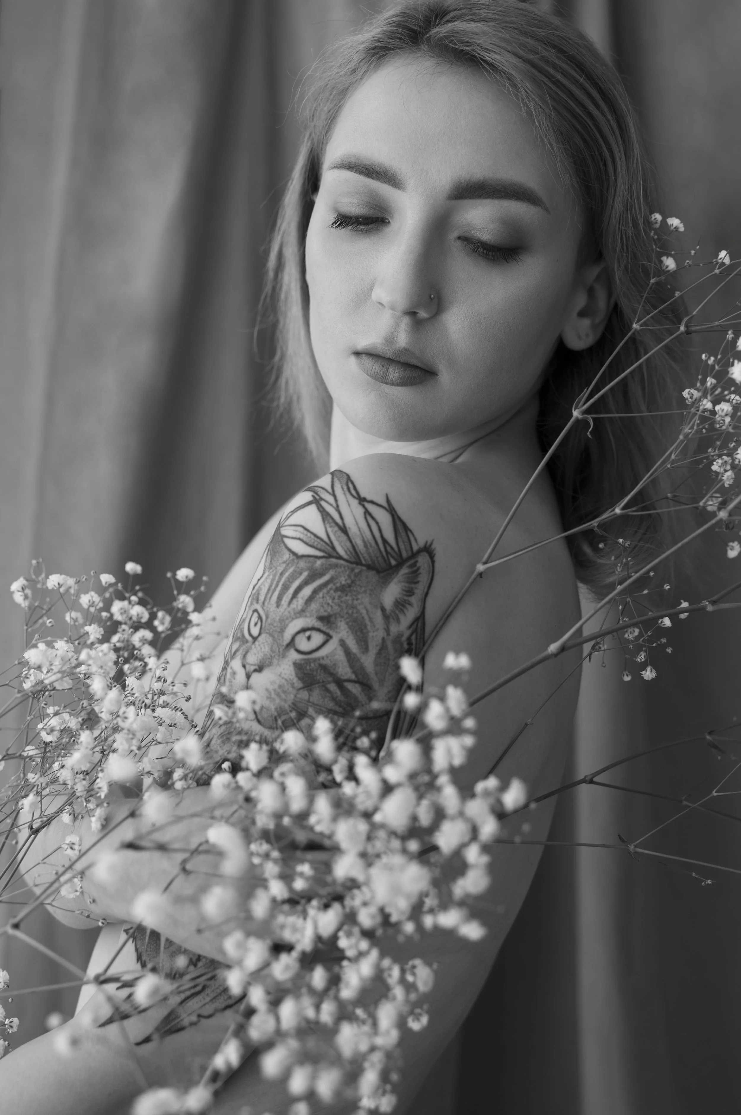 Black and white photo of a woman with closed eyes, media makeup, a nose piercing, and a floral tattoo on her shoulder, holding and surrounded by small flowers.