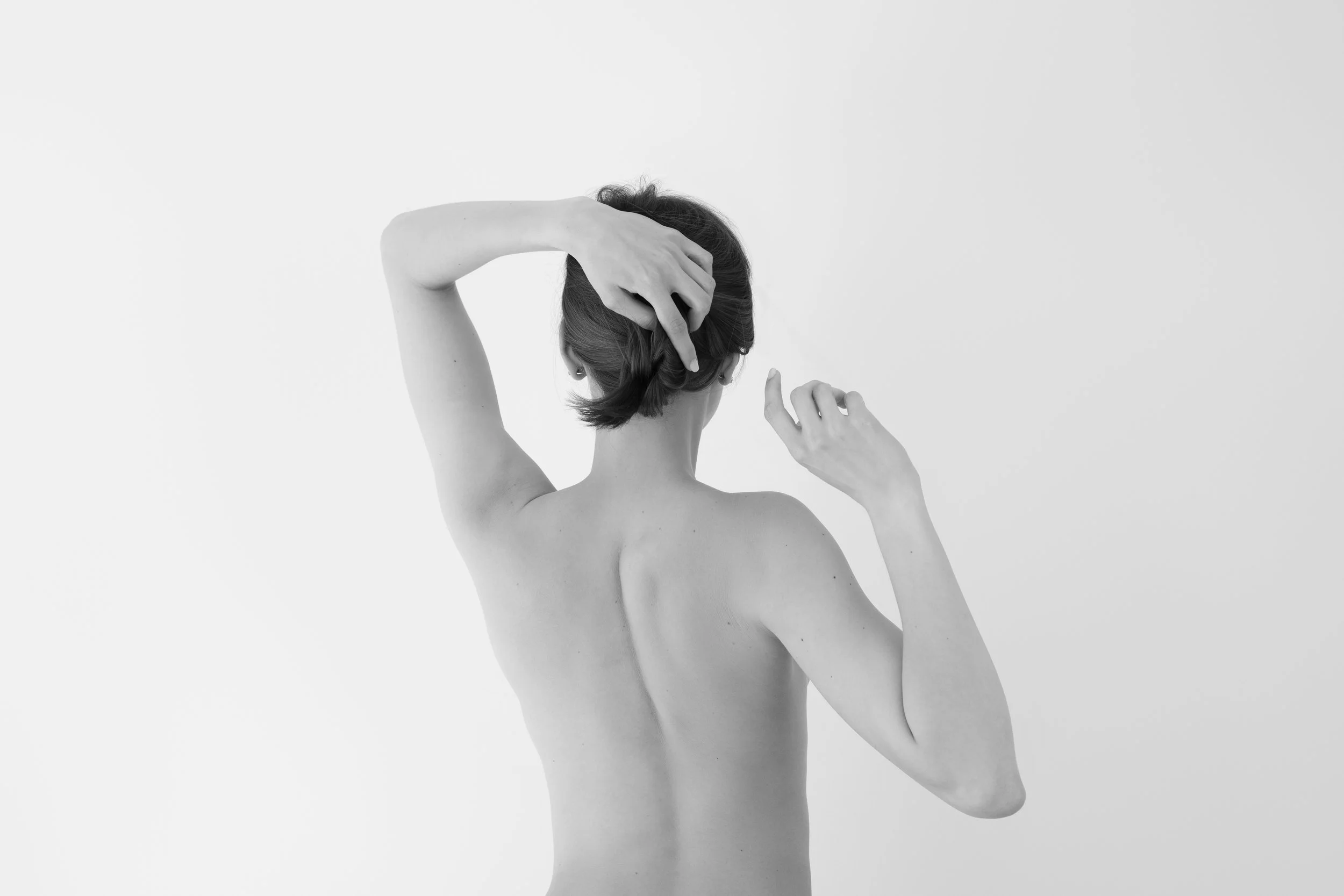 A black and white photo of a woman with short hair, topless, with her back turned, her left arm raised, and her hand touching her head.