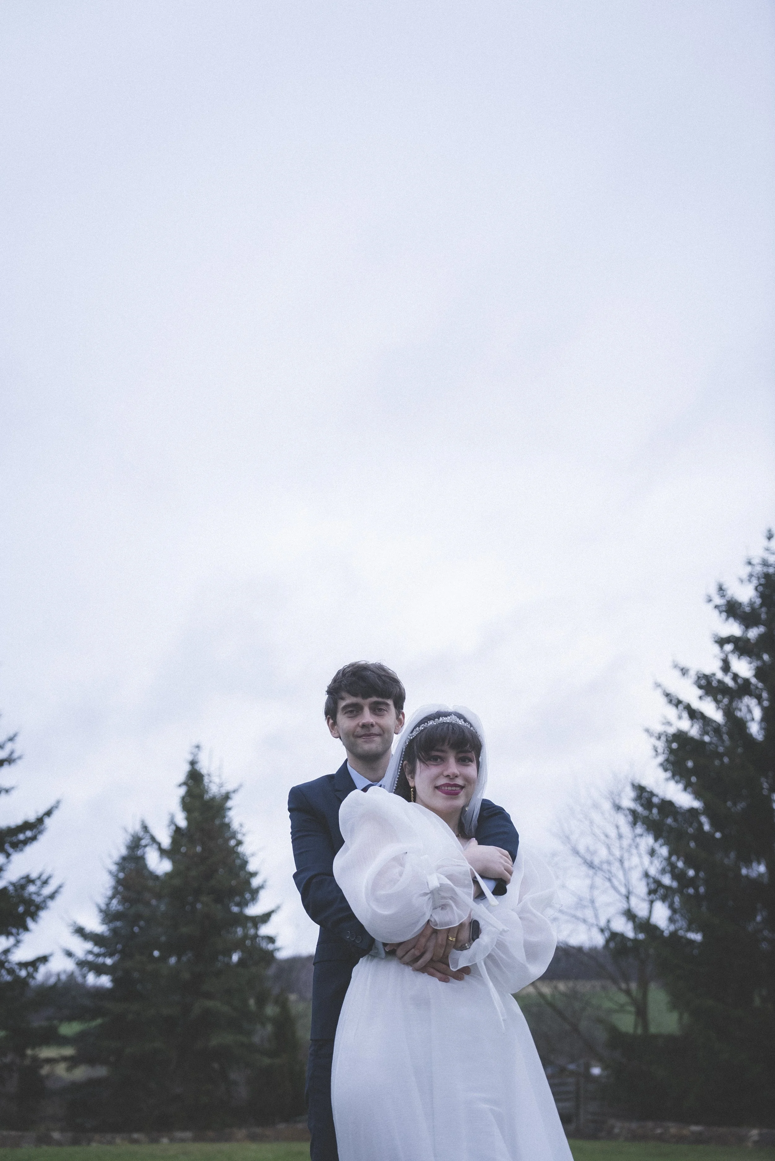 A newlywed couple standing outdoors, with the groom in a suit and the bride in a white dress and veil, surrounded by trees and an overcast sky.