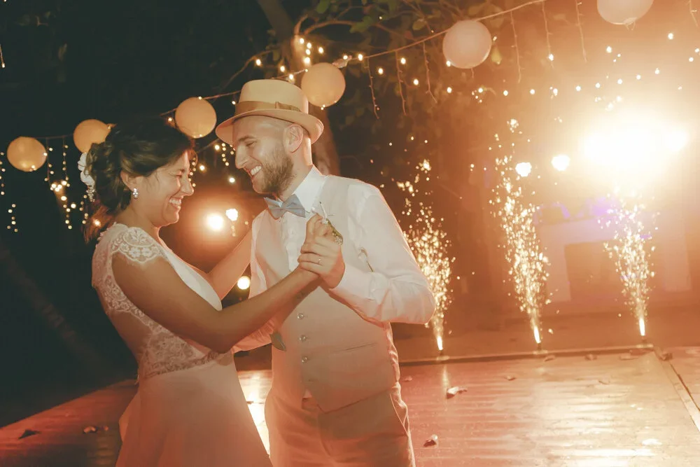 A bride and groom dancing at their wedding reception at night with string lights and fireworks in the background.