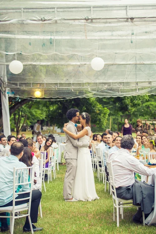 A bride and groom share a dance during their outdoor wedding reception, surrounded by seated guests under a canopy, with green trees in the background.