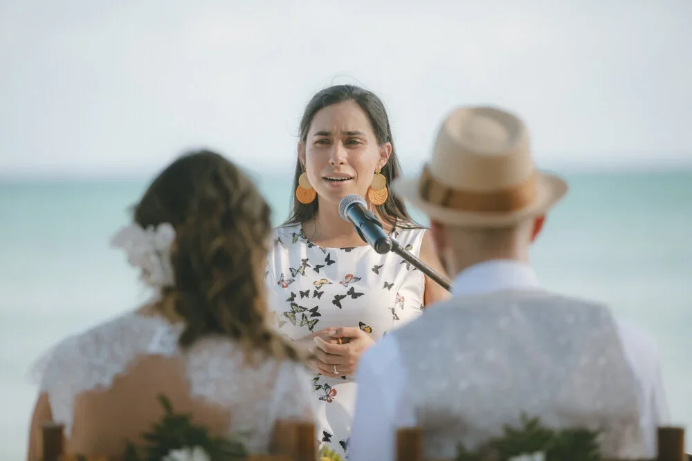 A woman gives a speech or vows at a beach wedding ceremony, with the ocean in the background. She is speaking into a microphone, wearing floral earrings and a dress with butterfly patterns. A man wearing a hat and a woman with flowers in her hair sit facing her.