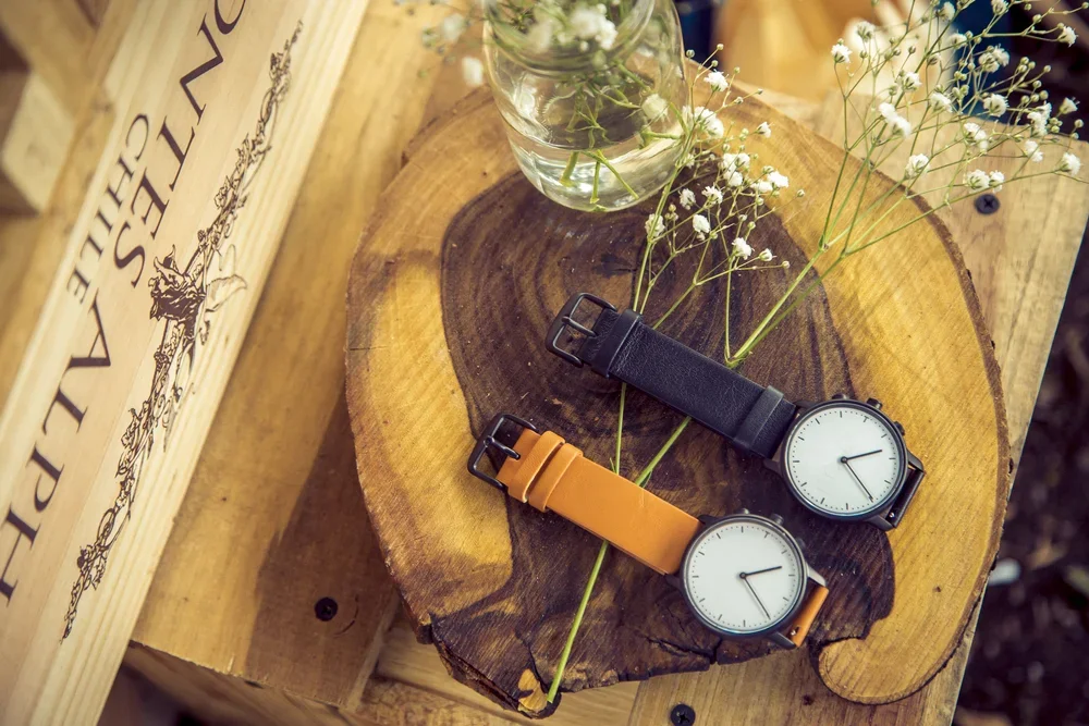 Two minimalist wristwatches with leather straps, one black and one orange, are placed on a rustic wooden surface with small white flowers and a glass jar.