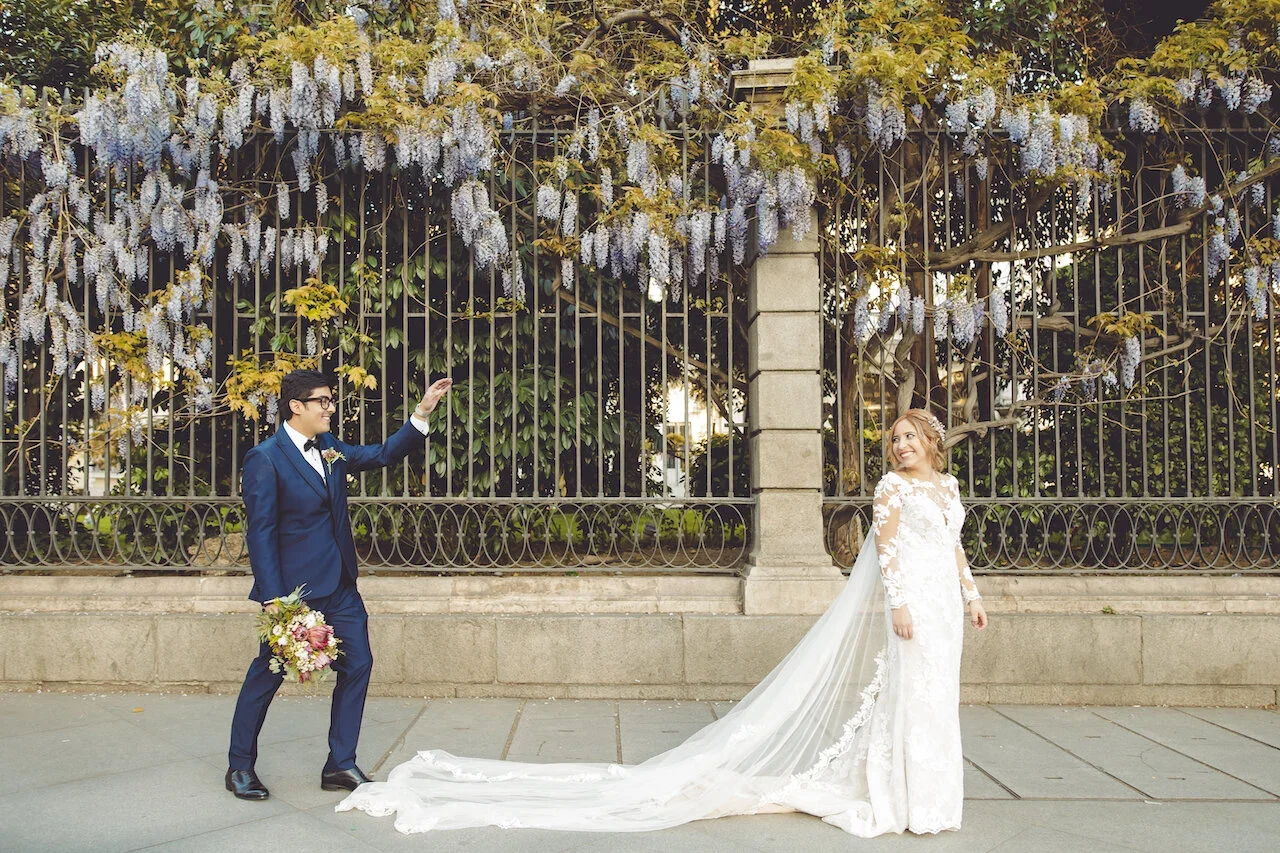A bride in a white lace wedding dress with a long train and veil smiling at a groom in a navy suit holding a bouquet of flowers, standing outdoors in front of a decorative metal fence with climbing purple wisteria flowers.