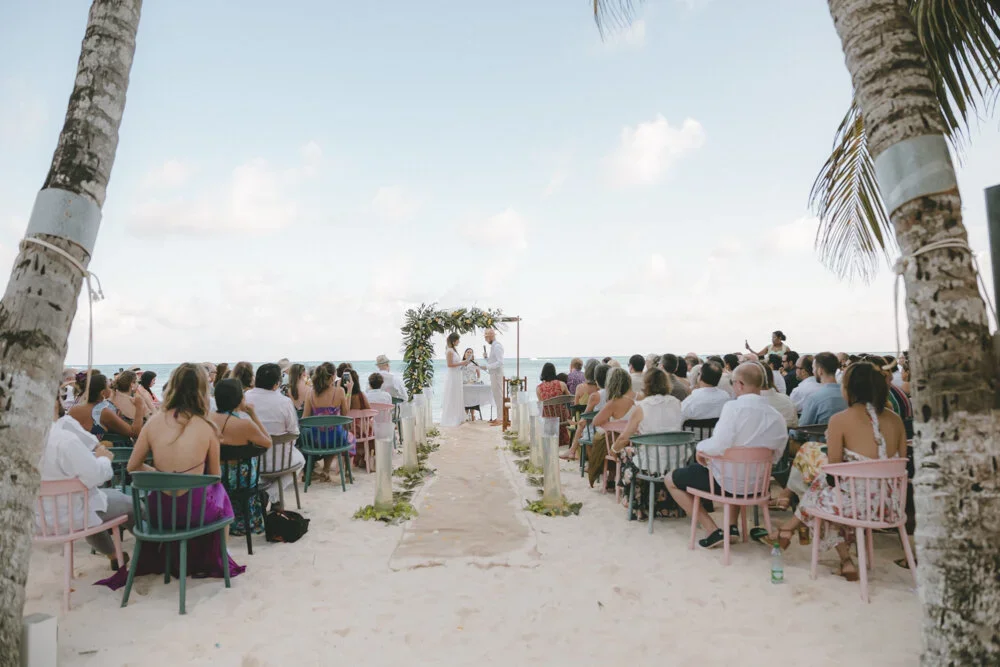 Outdoor beach wedding ceremony with guests seated on colorful chairs, officiant and couple standing under a floral arch, ocean in the background, palm trees framing the scene.