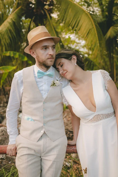 A couple dressed in wedding attire stands close together outdoors surrounded by lush greenery, including large palm leaves. The man wears a beige vest, white shirt, turquoise bow tie, and a beige hat, while the woman wears a white dress with lace details and rests her head on his shoulder.