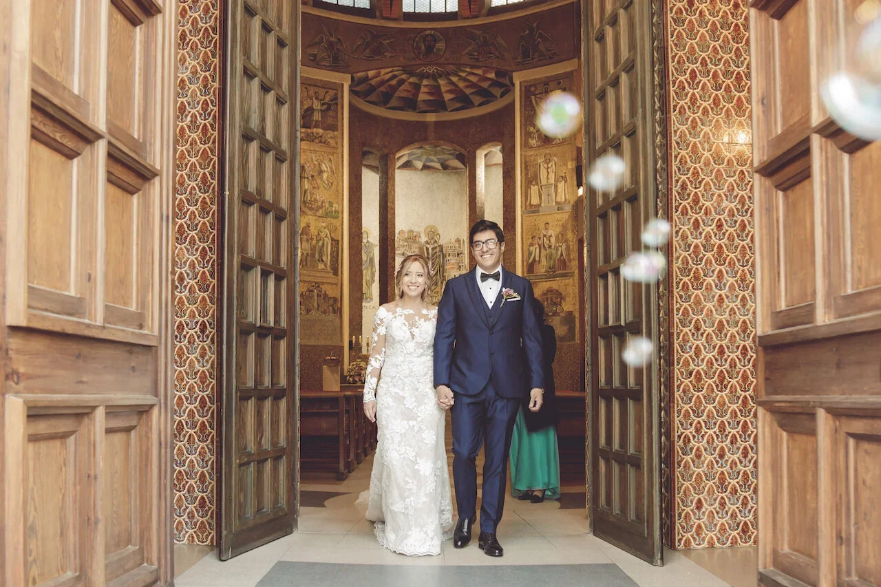A bride and groom walking into a church, holding hands and smiling, with a man in the background. The bride is in a white lace wedding dress and the groom is in a navy blue suit with a bow tie. The church has wooden doors and ornate paintings on the walls.