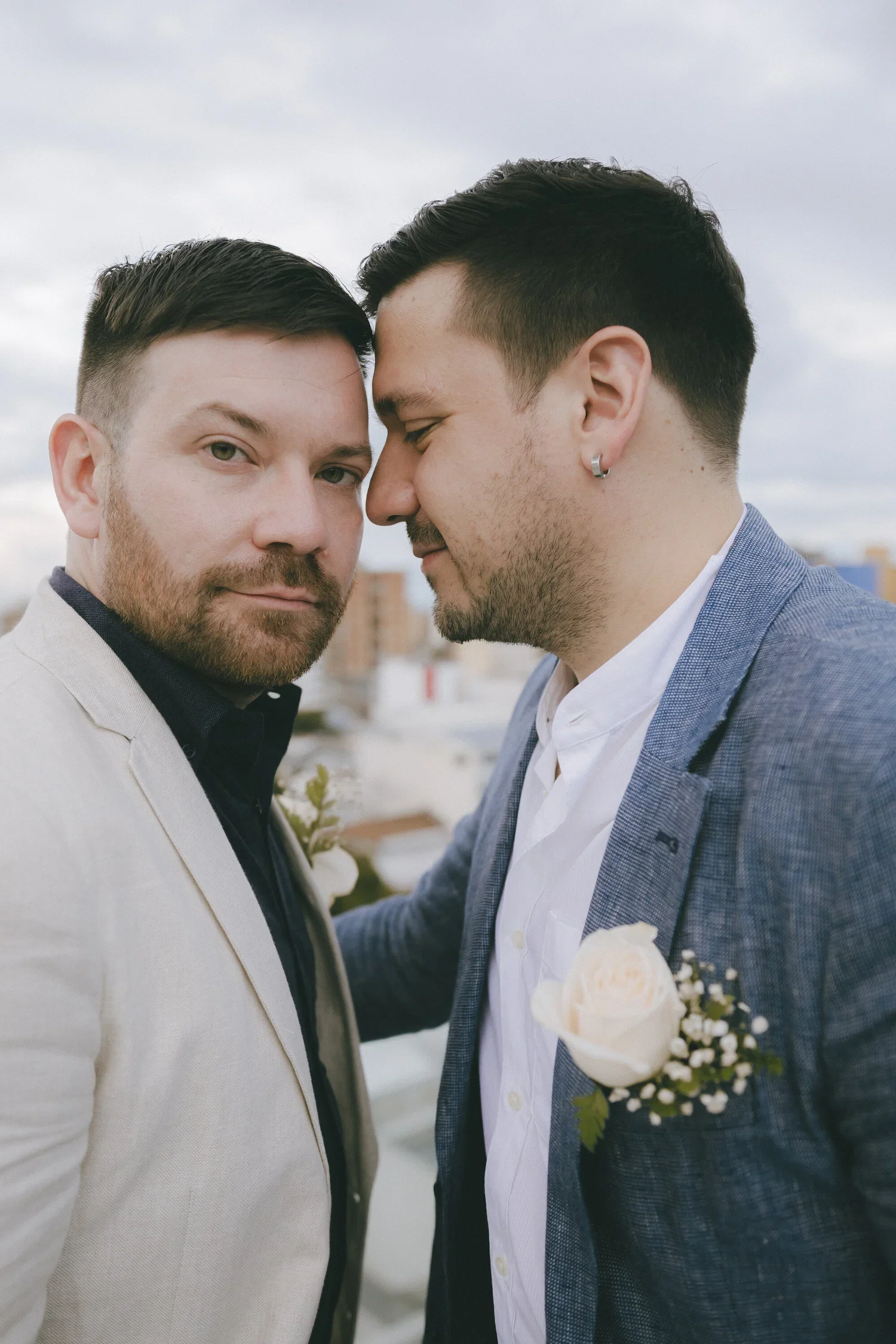 Two men stand close together with their foreheads touching, outdoors with cloudy sky, both dressed in suits with flowers pinned on their lapels, suggesting a wedding or romantic moment.