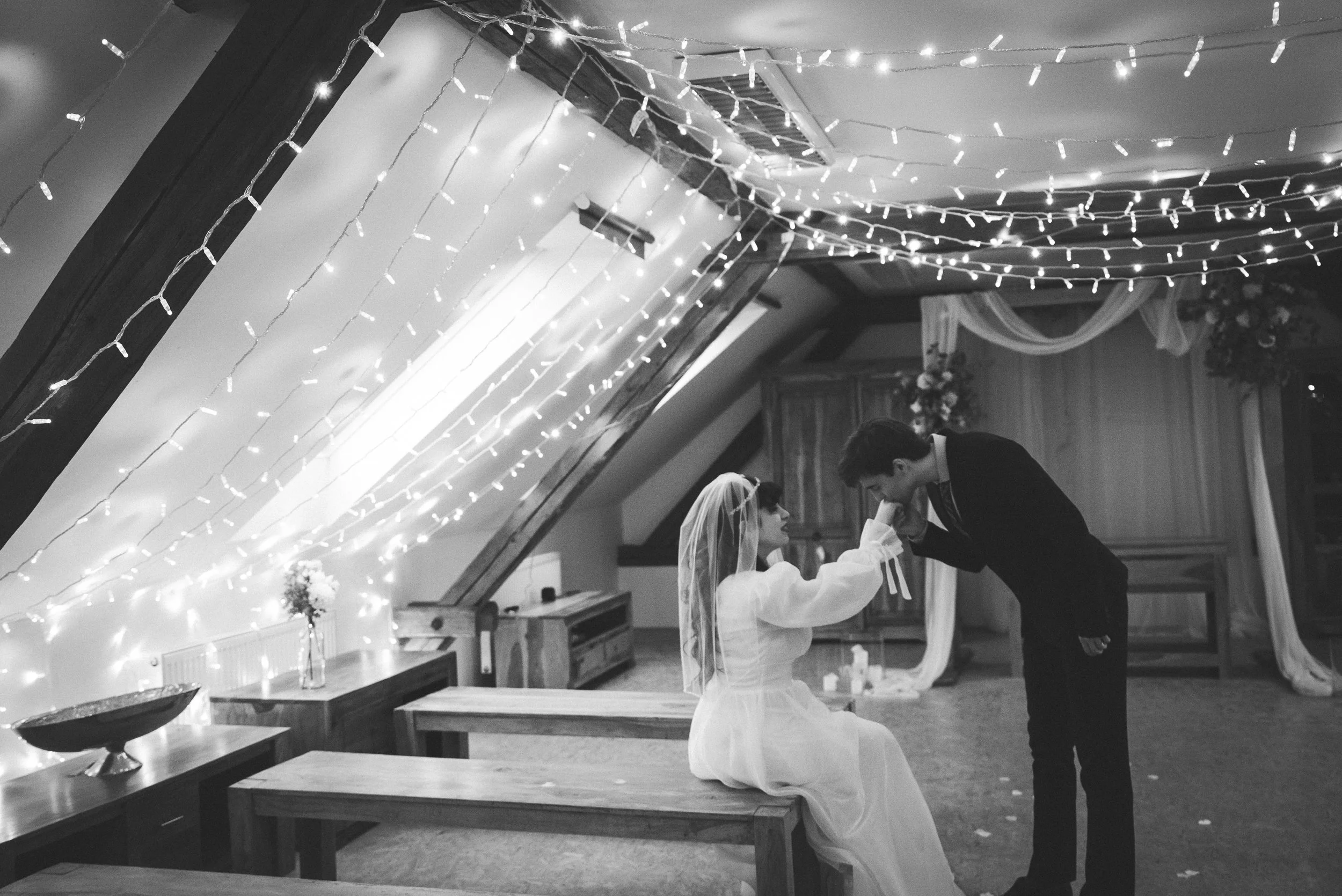 A black and white photo of a bride and groom during their wedding ceremony in a decorated venue with string lights hanging from the ceiling. The groom is leaning towards the bride, holding her hand. The bride is sitting on a bench, wearing a wedding dress and veil, while the groom is dressed in a suit.
