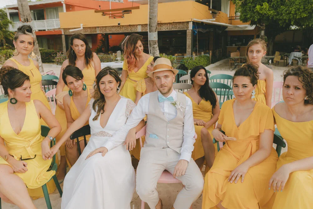 A group of women with light skin in yellow dresses and one man with light skin in a beige vest, white shirt, and straw hat, sitting and standing around during a daytime outdoor wedding on the beach.