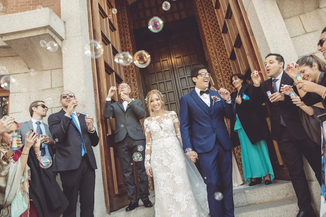 Bride and groom exiting a church holding hands, surrounded by friends and family blowing bubbles.