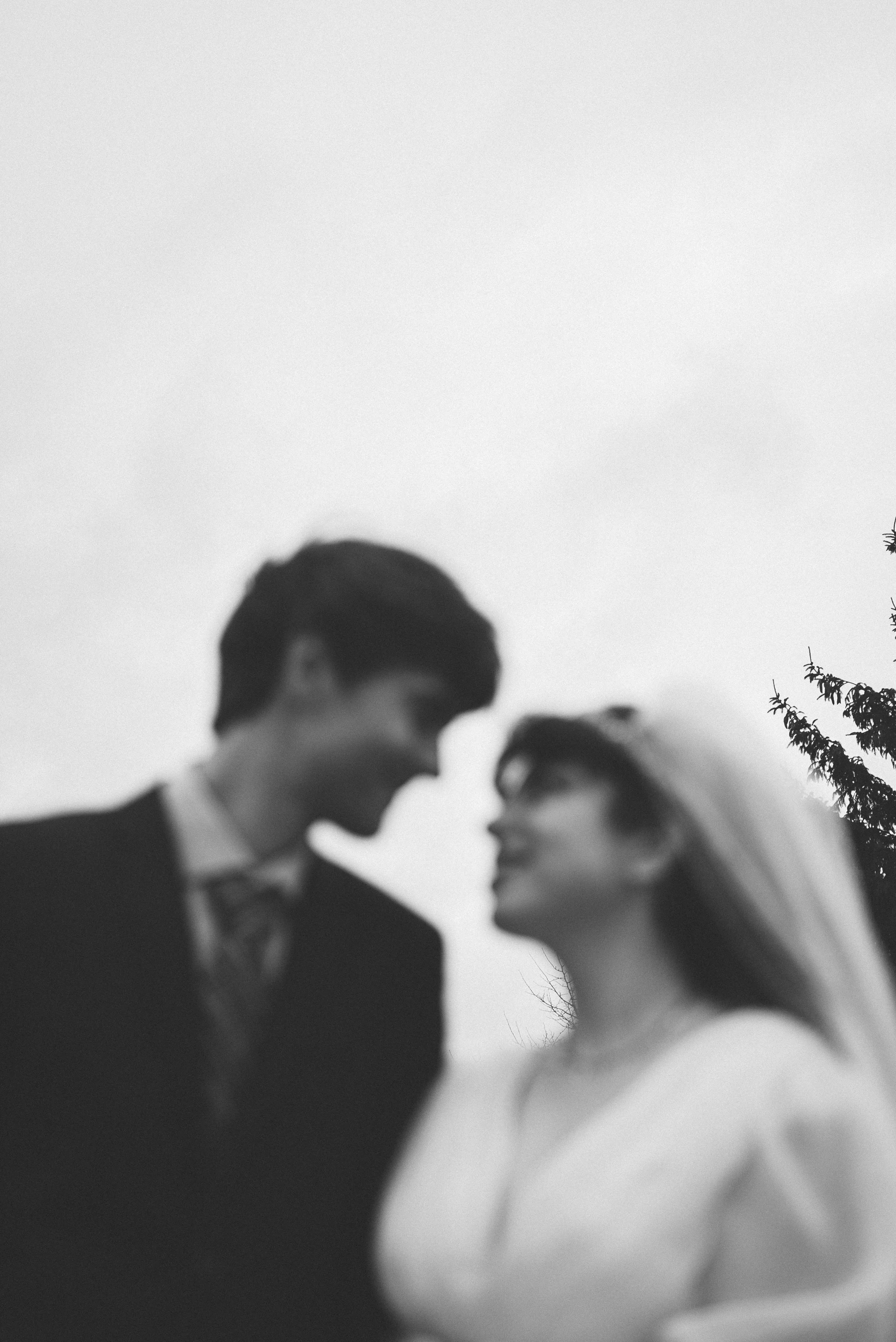 Black and white photo of a man and woman in wedding attire, close to each other, outdoors, with trees in the background.