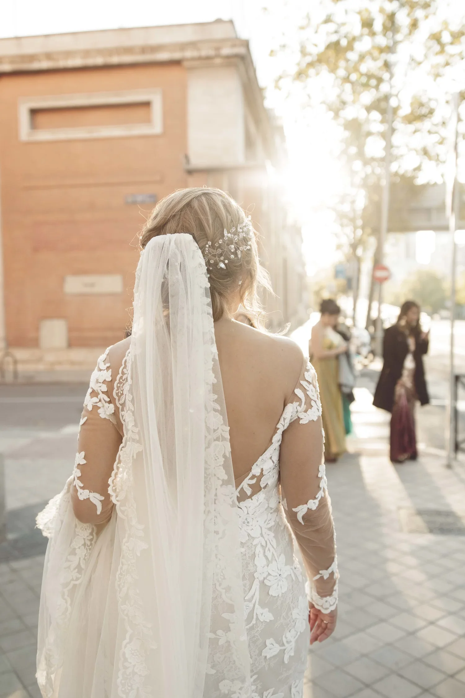 Bride in a white lace wedding dress with floral embroidery and a veil, standing outdoors during sunset, with people in the background.