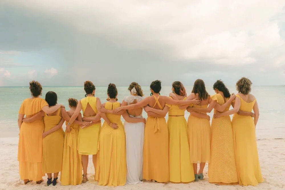 Group of women in yellow dresses standing on a beach with ocean and cloudy sky in background, facing away from the camera, embracing each other.