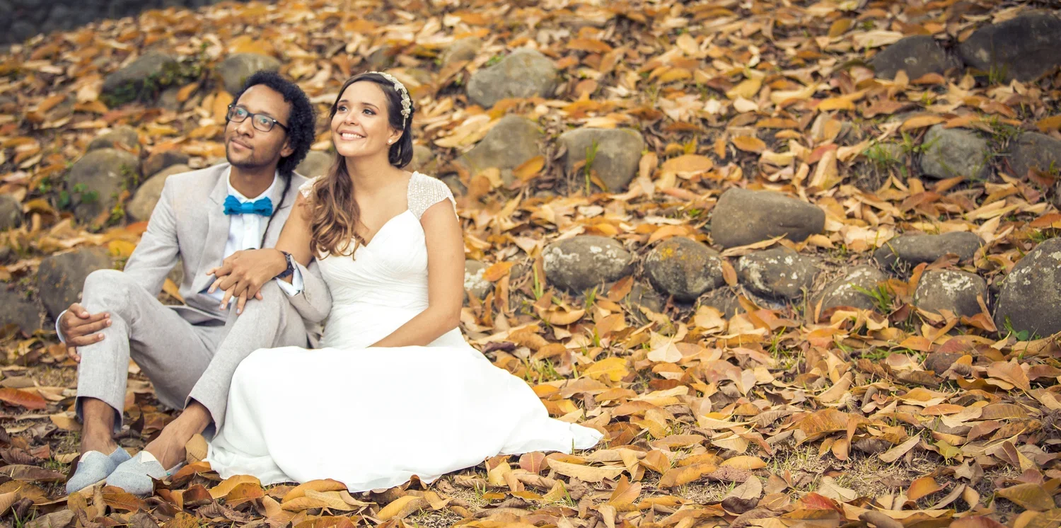 A couple in wedding attire sitting on the ground covered with fall leaves, with a backdrop of rocks and foliage.