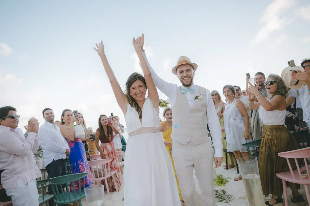 Bride and groom holding hands up together at their outdoor wedding celebration, surrounded by guests clapping and cheering.