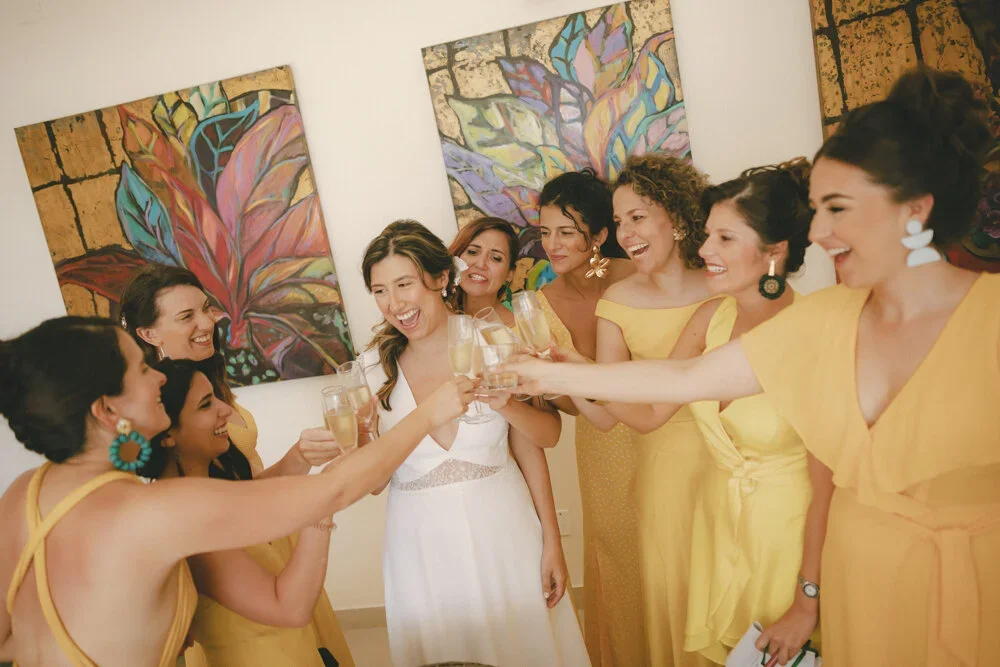 A bride in a white dress is surrounded by her bridesmaids in yellow dresses, celebrating with champagne in a room with colorful abstract art on the walls.