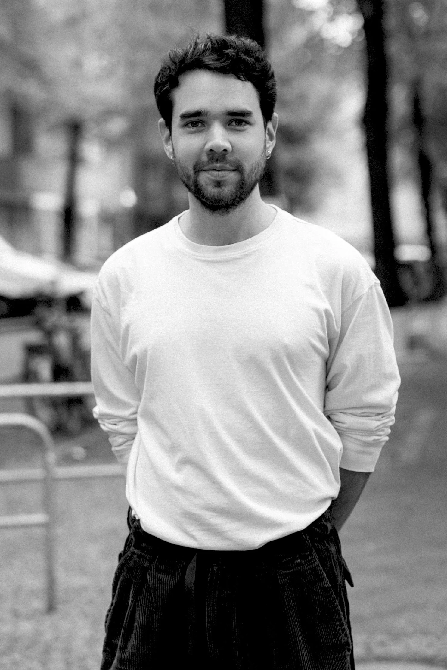 Black and white portrait of a young man with dark hair and a beard, wearing a plain white long-sleeve shirt, standing outdoors with blurred trees and chairs in the background.