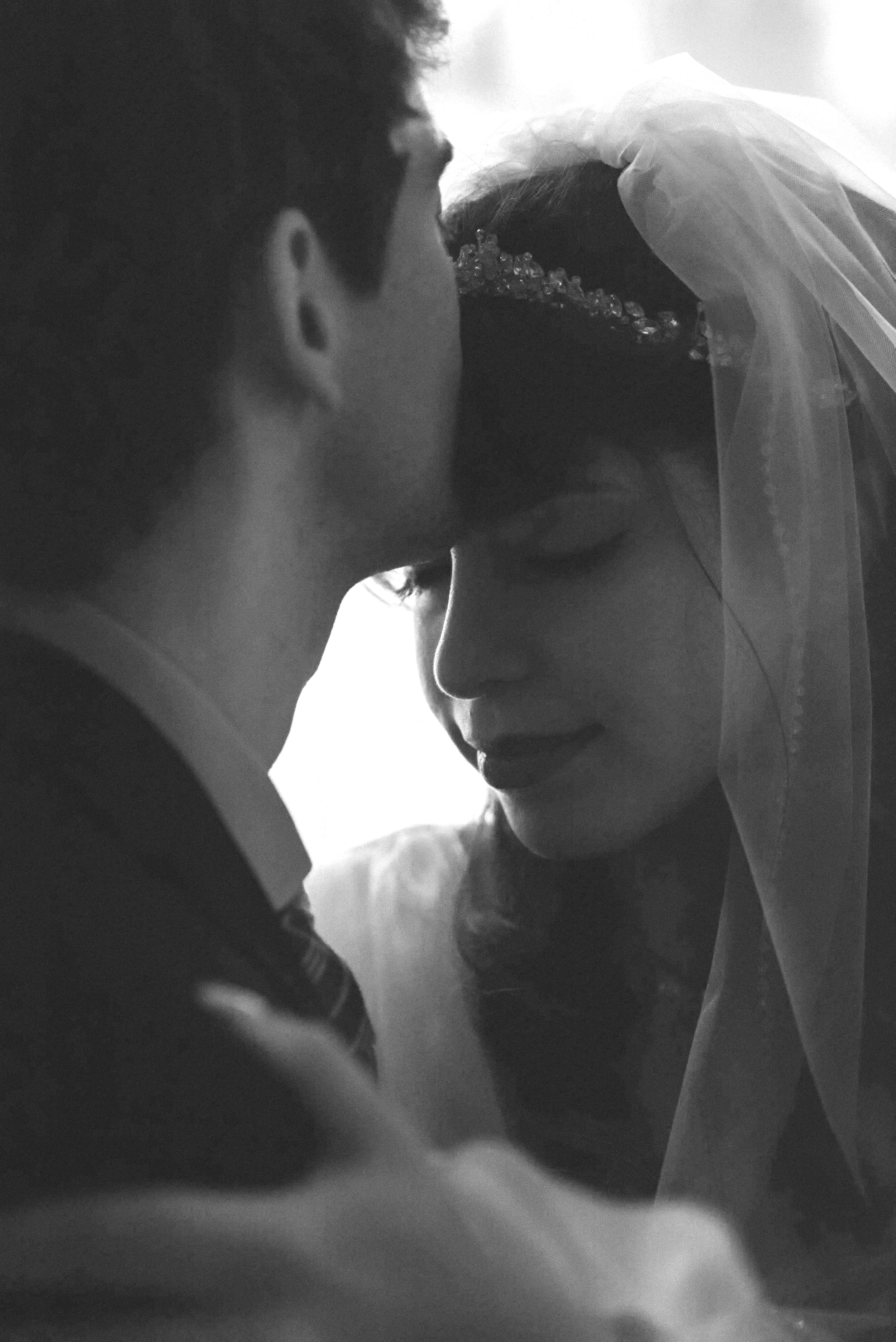 A black-and-white photograph of a bride and groom in an intimate moment, close together with their foreheads and noses touching. The bride is wearing a wedding veil and has a jeweled hairpiece, while the groom is in a dark suit, both capturing a tender, emotional connection.