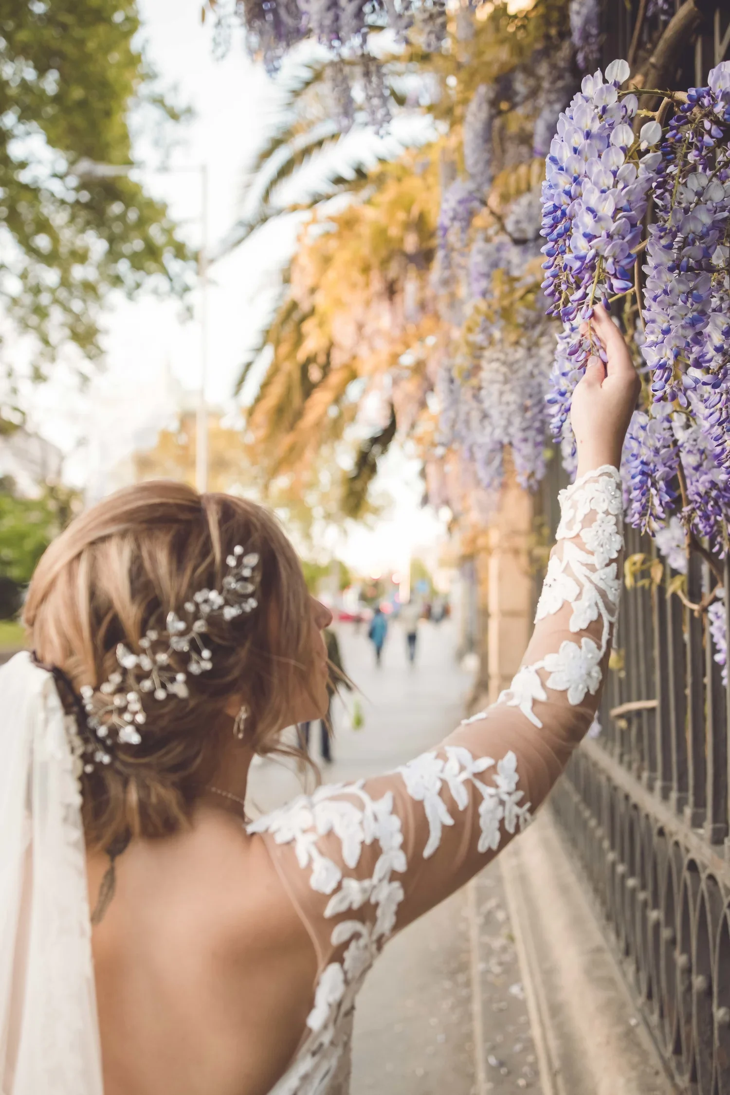 A woman in a white lace wedding dress is reaching up to touch purple wisteria flowers growing on a fence along a city street.