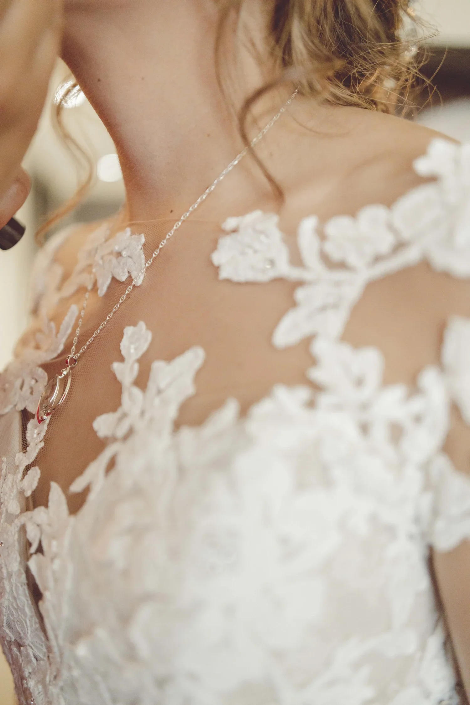 Close-up of a woman wearing a wedding dress with lace and floral details, showing her shoulder and the back of her neck, with curly hair and a delicate necklace.