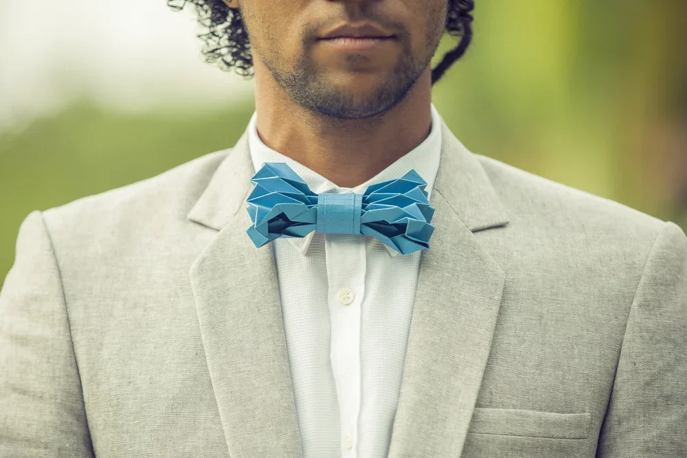 Close-up of a man wearing a light grey suit and a white shirt, with a blue origami-style bow tie, outdoors with blurred greenery in the background.