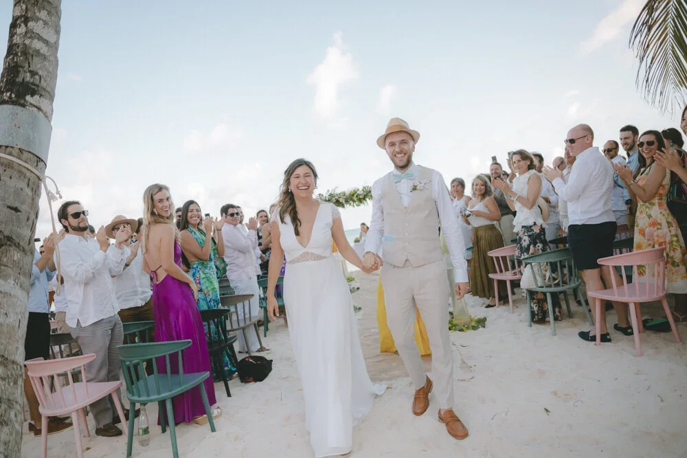 A bride and groom walking hand in hand down the aisle at a beach wedding, surrounded by guests clapping and smiling.