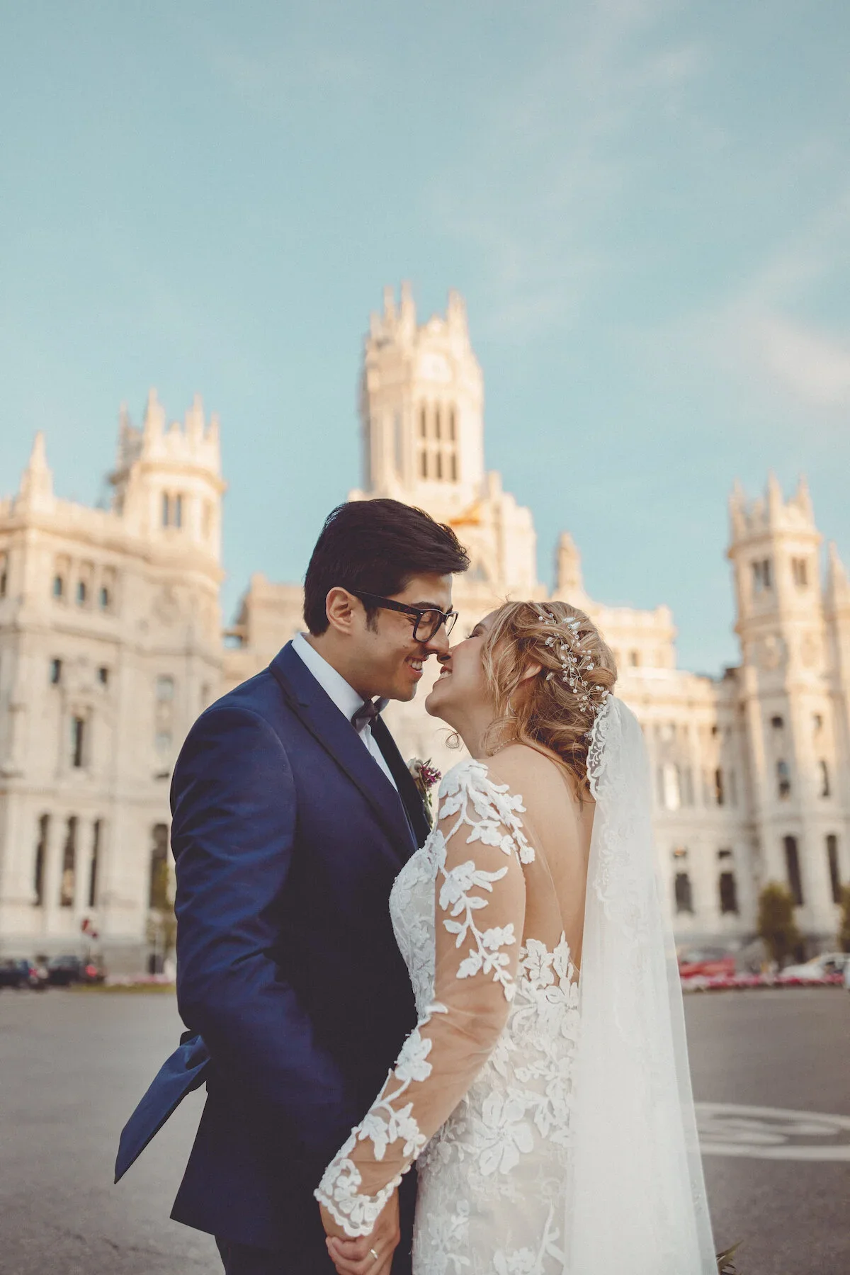 A bride and groom are standing close together, touching noses and smiling in front of a large castle-like building with towers, under a partly cloudy sky.